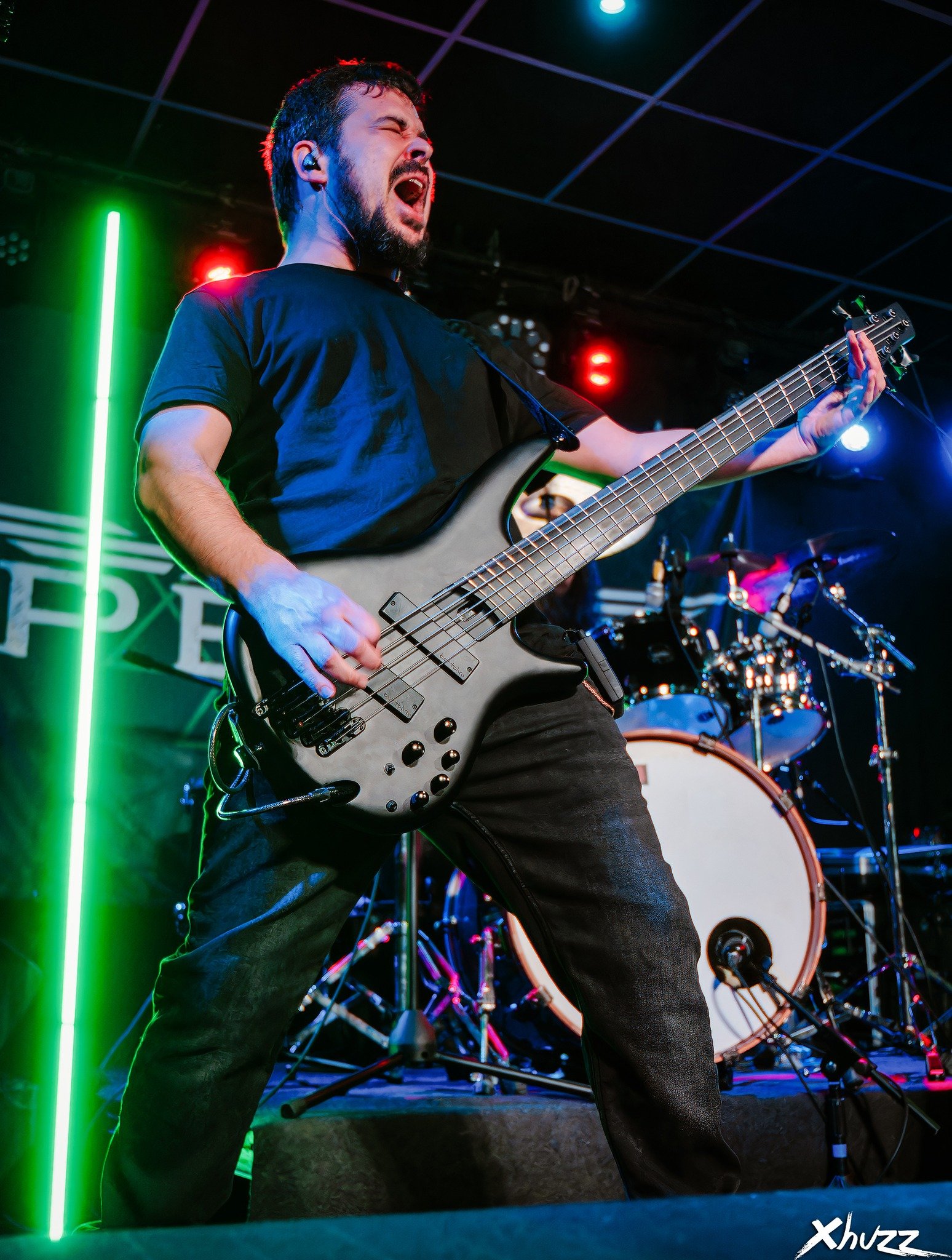 Un hombre tocando Metal moderno, groove metal, metalcore y deathcore en el bajo electrico en un escenario, con expresión apasionada, en un concierto con luces de colores y batería en el fondo.