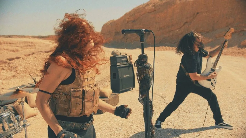 Dos músicos de la banda de madrid de Metal moderno, groove metal, metalcore y deathcore Rabia Perez tocando en un paisaje desértico, uno con cabello pelirrojo y suelto, y otro con cabello largo y oscuro, tocando una guitarra eléctrica. Hay equipo de 