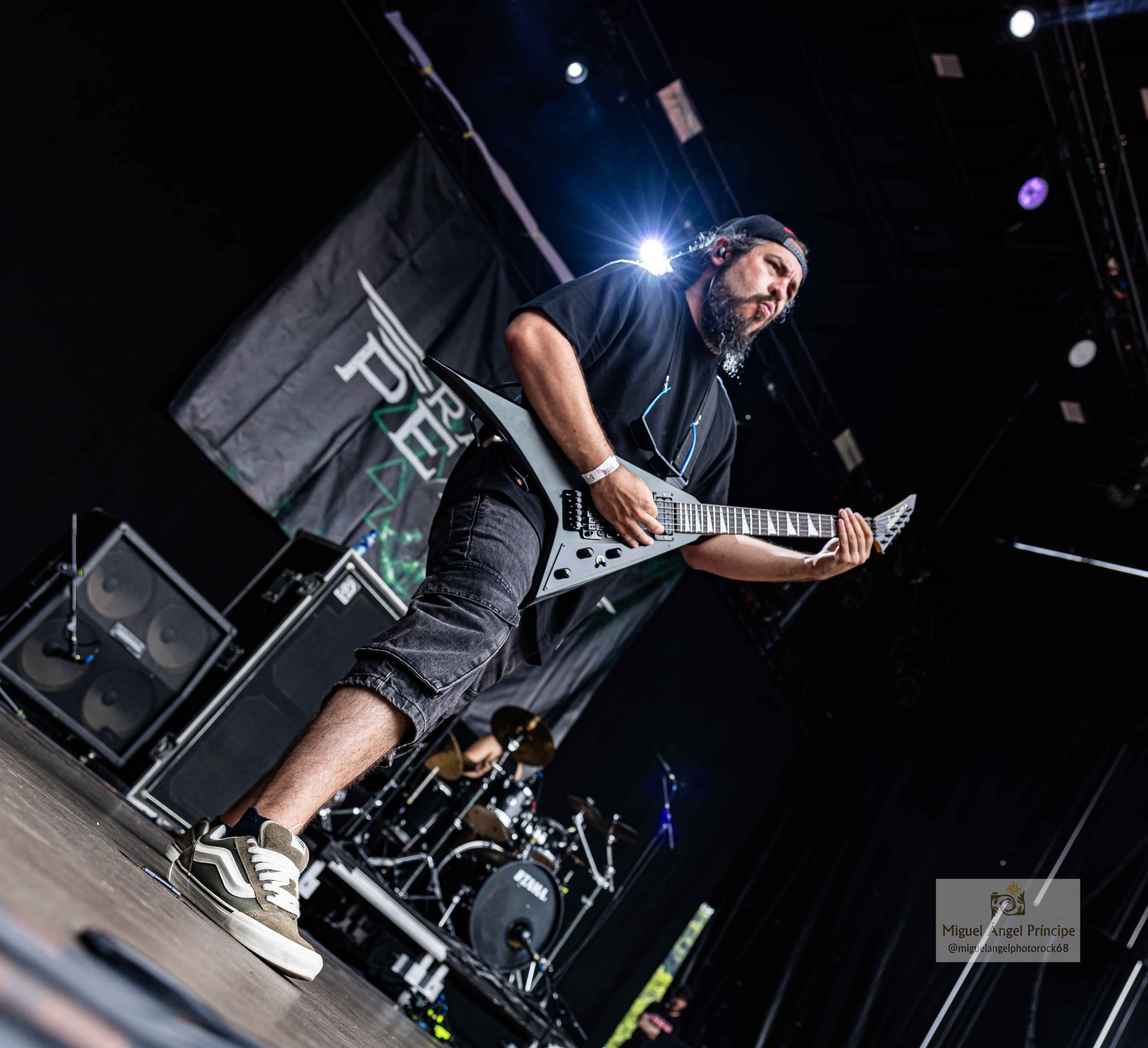 Un hombre fundador de la banda de Metal moderno, groove metal, metalcore y deathcore de Madrid tocando guitarra eléctrica en un escenario durante un concierto, con una bandera en el fondo, usa camiseta negra, pantalones cortos y zapatillas deportivas