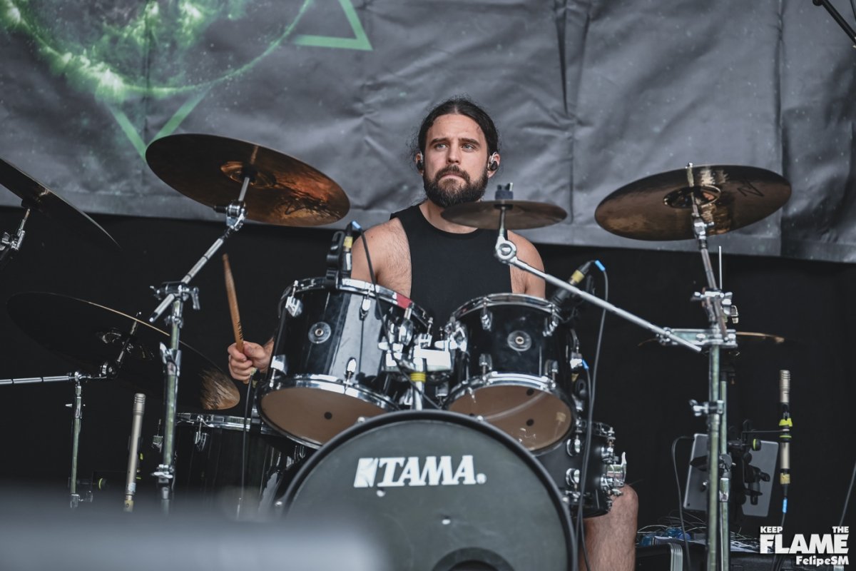 Un hombre con barba toca Metal moderno, groove metal, metalcore y deathcore en la batería en un escenario, lleva una camiseta sin mangas y auriculares.