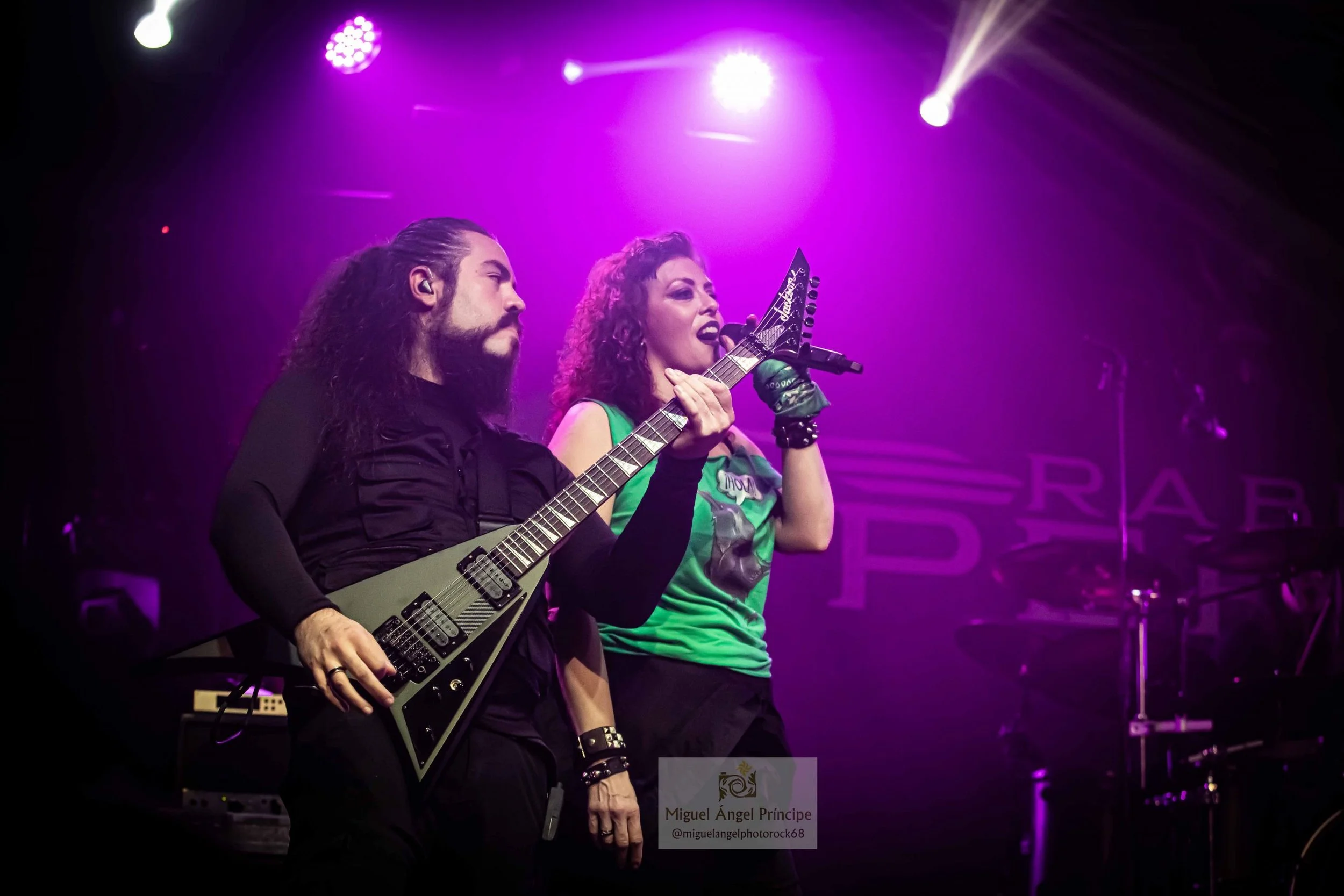 Dos músicos en escenario de Madrid con luces moradas y blancas, uno tocando guitarra eléctrica y la otra cantando con micrófono, en un concierto de Metal moderno, groove metal, metalcore y deathcore.
