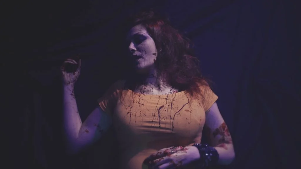 Mujer con expresión dramática, vestida con camiseta naranja, en un ambiente oscuro con efectos de sangre en su rostro y ropa.
