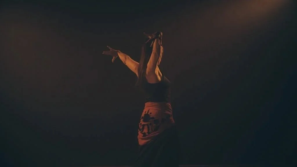 Una bailarina en un escenario oscuro, con brazos extendidos y vestida con ropa negra y un delantal anaranjado que tiene un patrón.