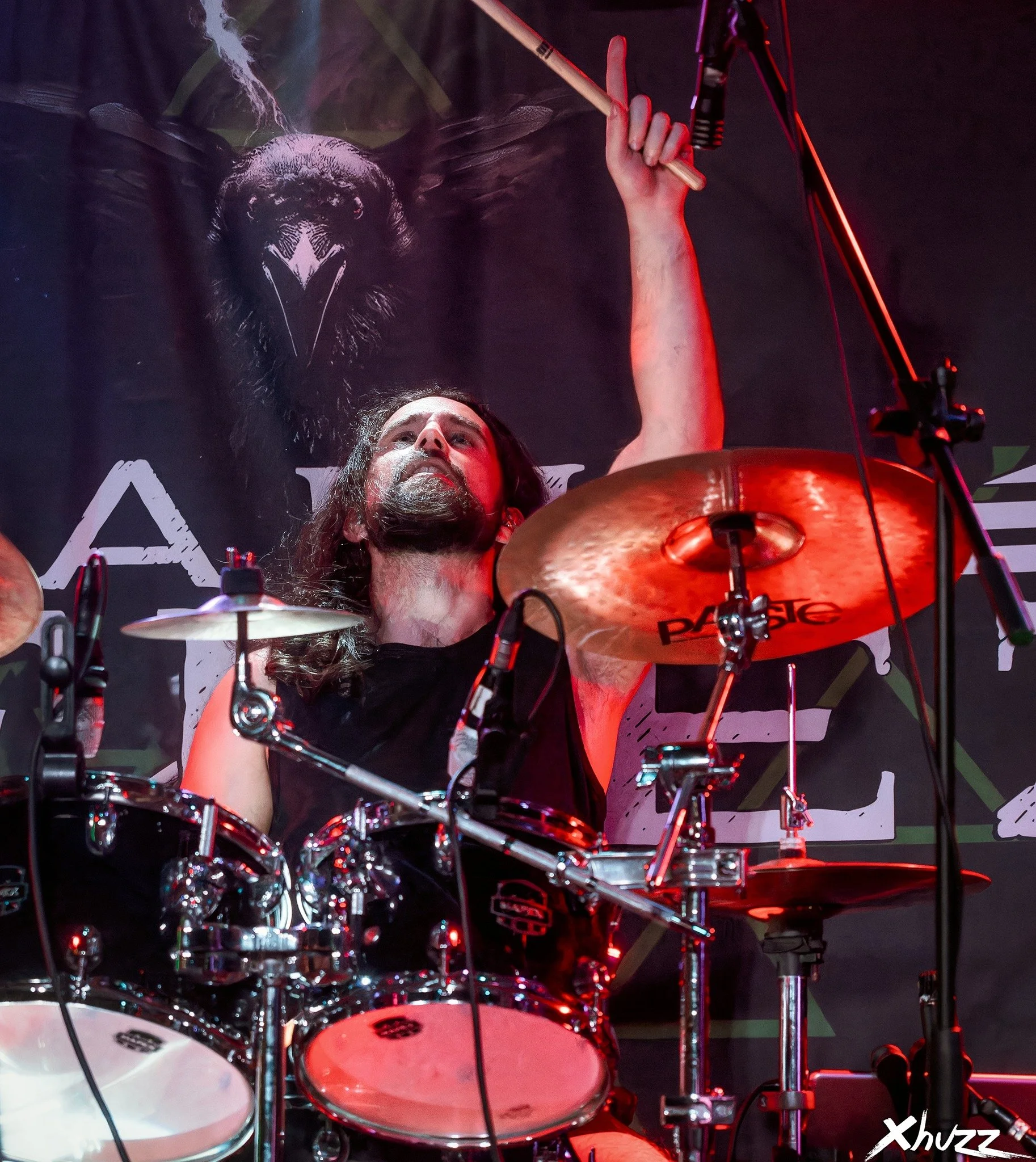 Baterista en concierto de la banda de Metal moderno, groove metal, metalcore y deathcore Rabia Perez con fondo oscuro y bandera con un animal y letras, iluminado por luces rojas, tocando su batería.