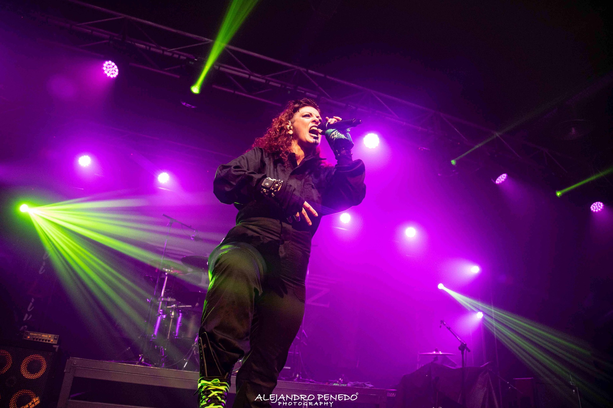 Una cantante de Metal moderno, groove metal, metalcore y deathcore con ropa negra, con cabello rizado, en medio de un escenario en Madrid con luces moradas y verdes, cantando en micrófono frente a un fondo de instrumentos musicales y equipo de sonido