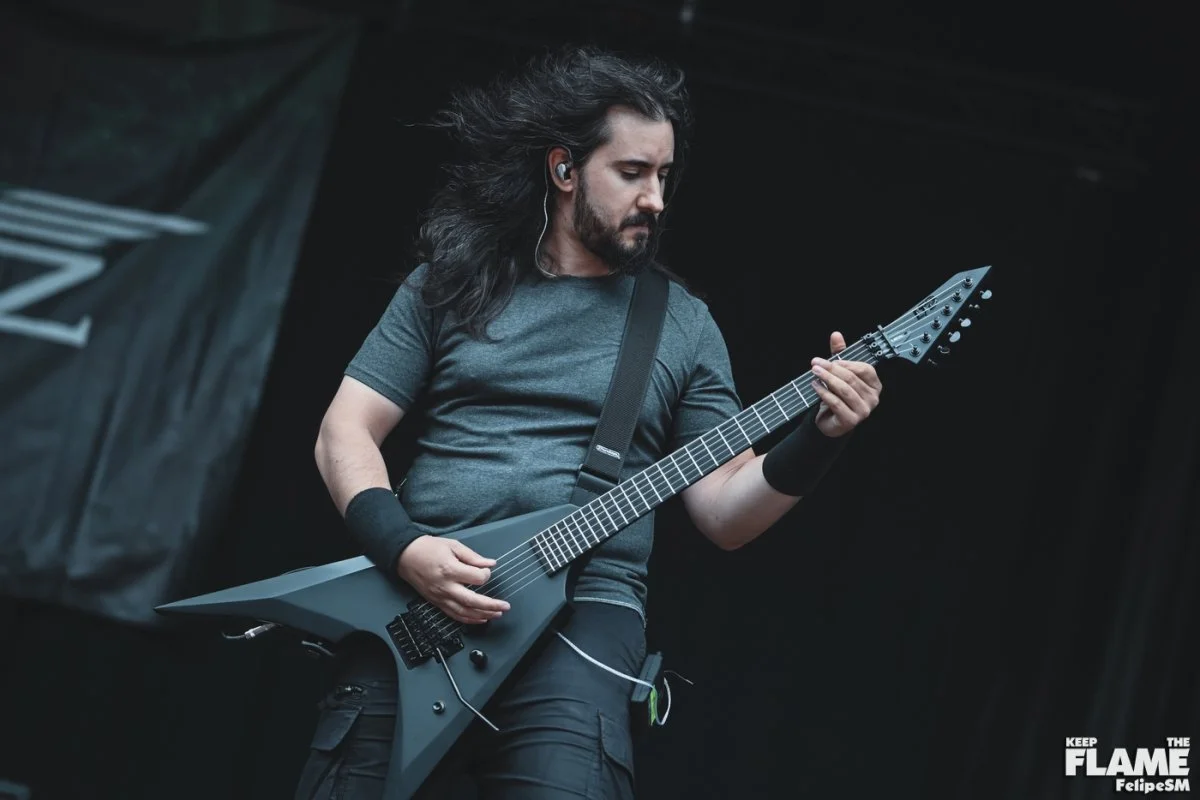 Hombre tocando Metal moderno, groove metal, metalcore y deathcore en guitarra eléctrica en un escenario, con cabello largo y enérgico, camiseta gris y auriculares y el telon de la banda Rabia Perez detrás