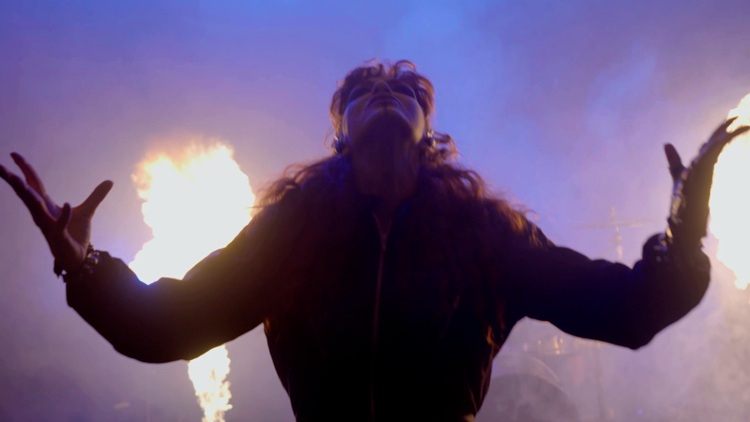 Mujer con cabello rizado y pelirojo con brazos extendidos, en medio de fuego y humo en un escenario oscuro en el rodaje del videoclip "los pájaros" de la banda de madrid de Metal moderno, groove metal, metalcore y deathcore Rabia Perez.