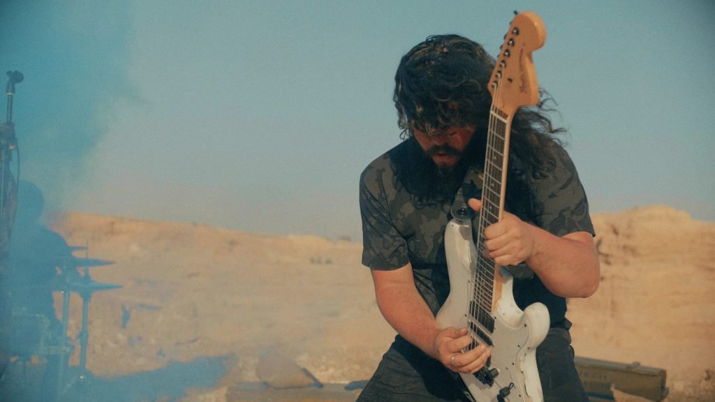 Gabriel Peñasco, guitarrista fundador de la banda de Metal moderno, groove metal, metalcore y deathcore Rabia Perez con cabello rizado y barba tocando una guitarra eléctrica en un entorno árido y despejado, con humo en el aire y un cielo claro.