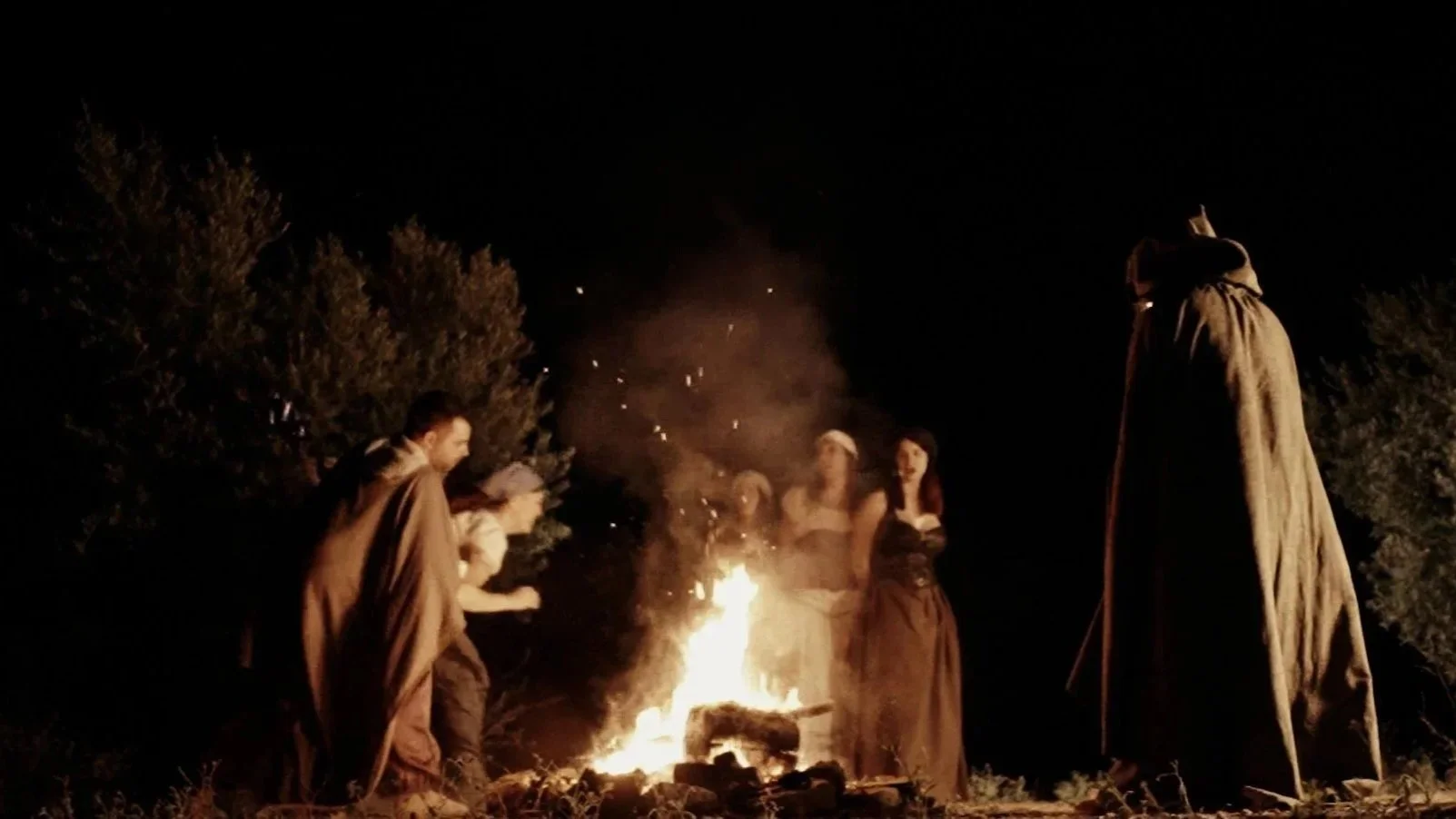 Grupo de personas alrededor de una fogata en la noche, algunas parecen estar contando historias o realizando una ceremonia al aire libre, con árboles en el fondo. rodajde videoclip de la banda de Metal moderno, groove metal, metalcore y deathcore.