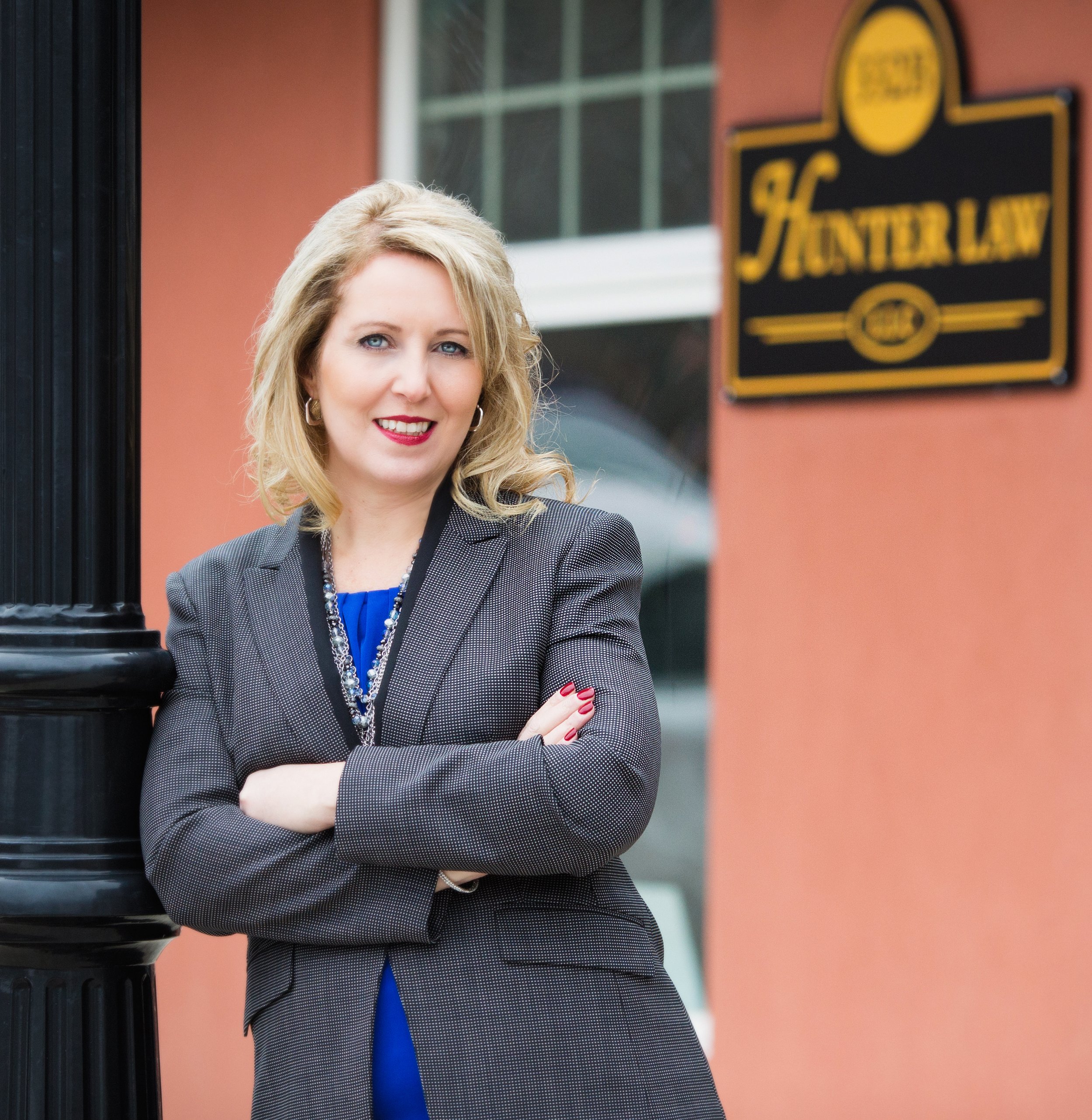 A female attorney in a gray blazer with a blue top, standing outside of her law firm with her arms crossed, in front of a black lamppost and a peach-colored building with a sign that reads 'HUNTER LAW, LLC'.