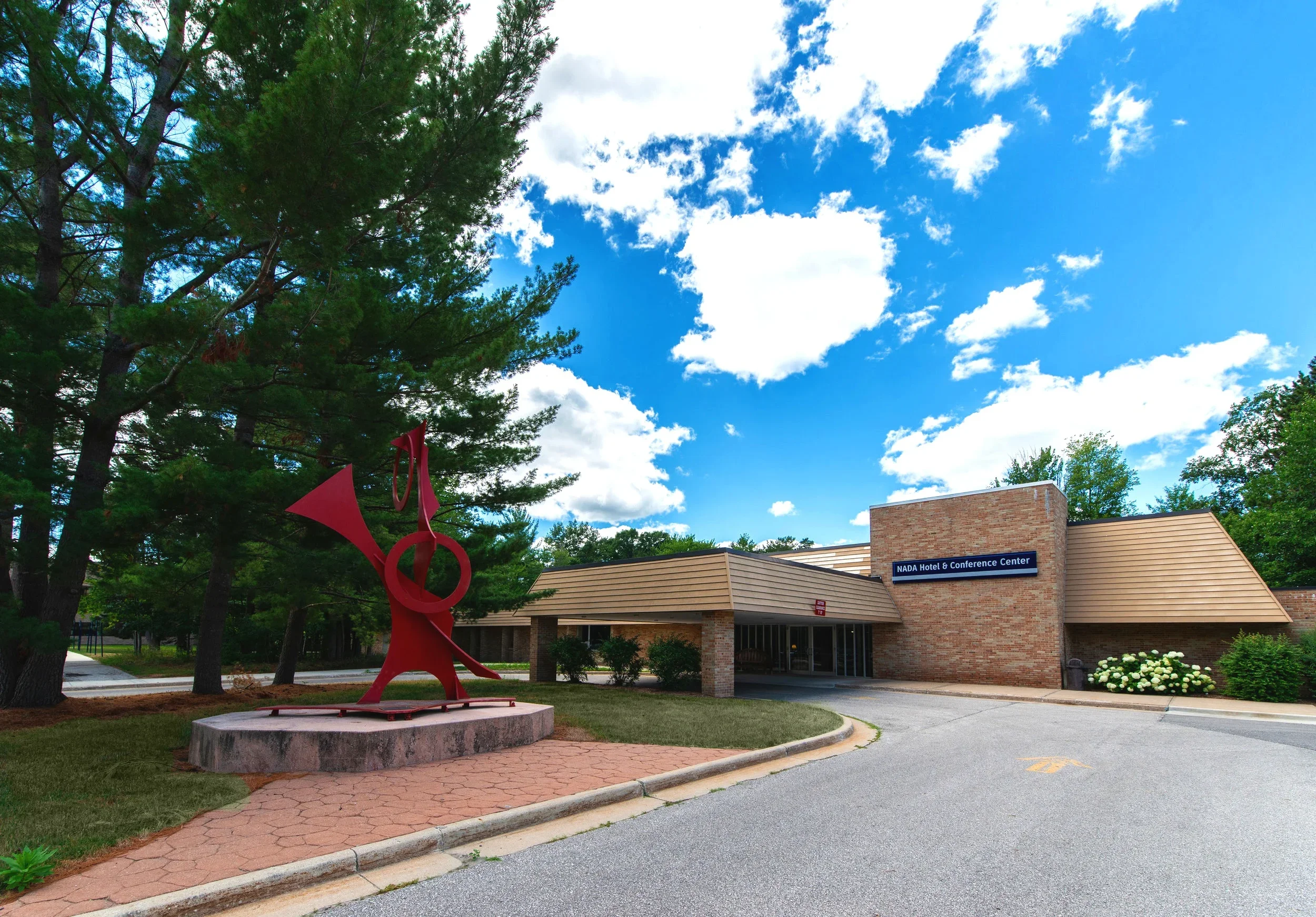 Exterior of NADA Hotel & Conference Center with a red abstract outdoor sculpture, trees, and a partly cloudy blue sky.