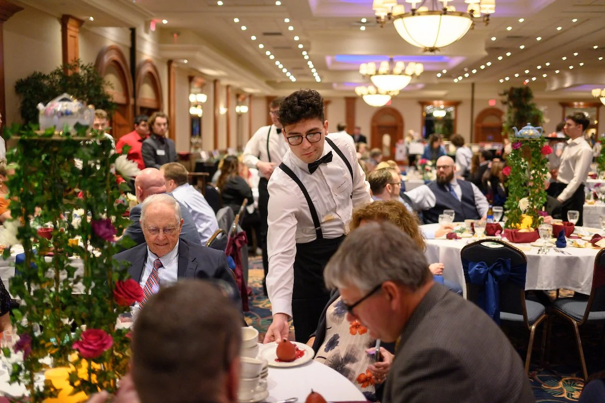 People dining at a banquet table in a decorated hotel ballroom with floral centerpieces, chandeliers, and colorful table settings.