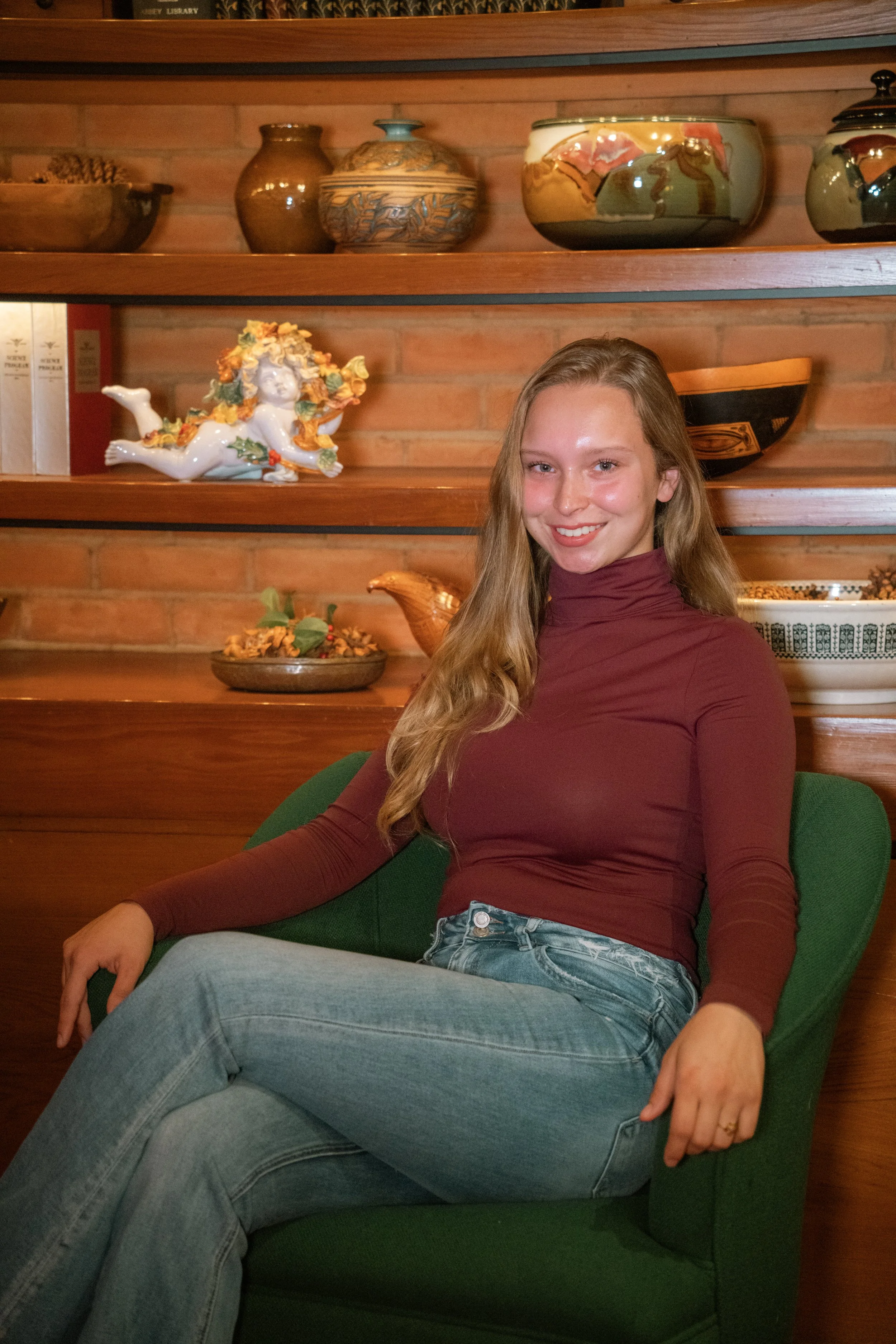 A young woman with long blonde hair, wearing a maroon long-sleeve shirt and blue jeans, sitting on a green chair in front of a wooden brick wall with decorative pottery and ceramics on shelves.