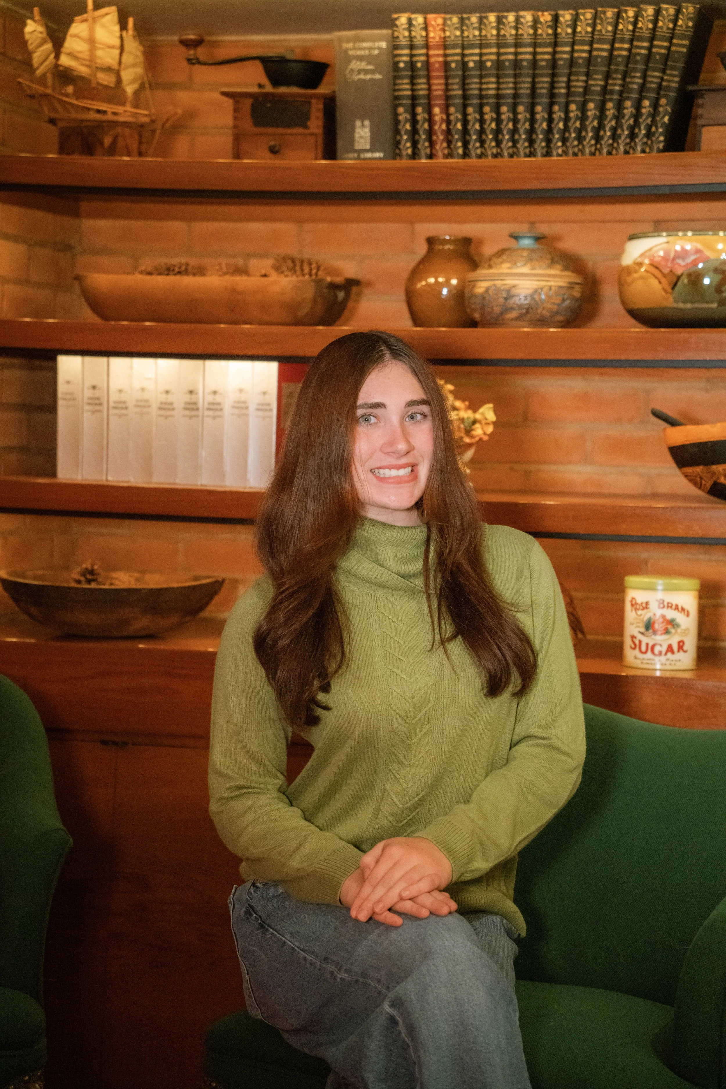 A woman with long brown hair wearing a light green sweater and gray jeans, sitting on a green chair with a wooden wall and shelves behind her. The shelves contain books, pottery, a bowl, and decorative items. There is a small container labeled "Sugar" on a middle shelf.