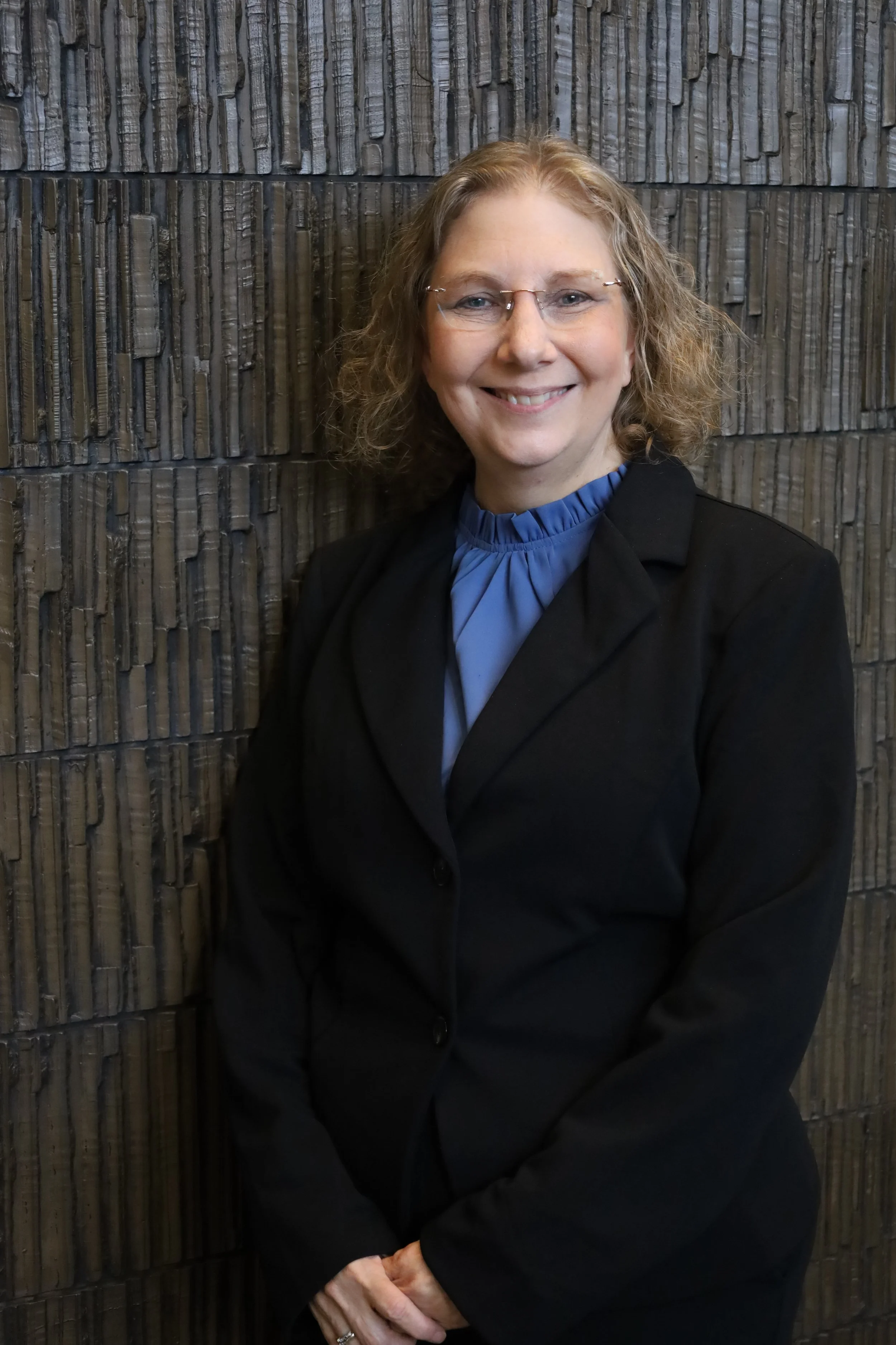 A woman with curly blonde hair, wearing glasses, a black blazer, and a blue blouse, standing in front of a textured brick wall, smiling.