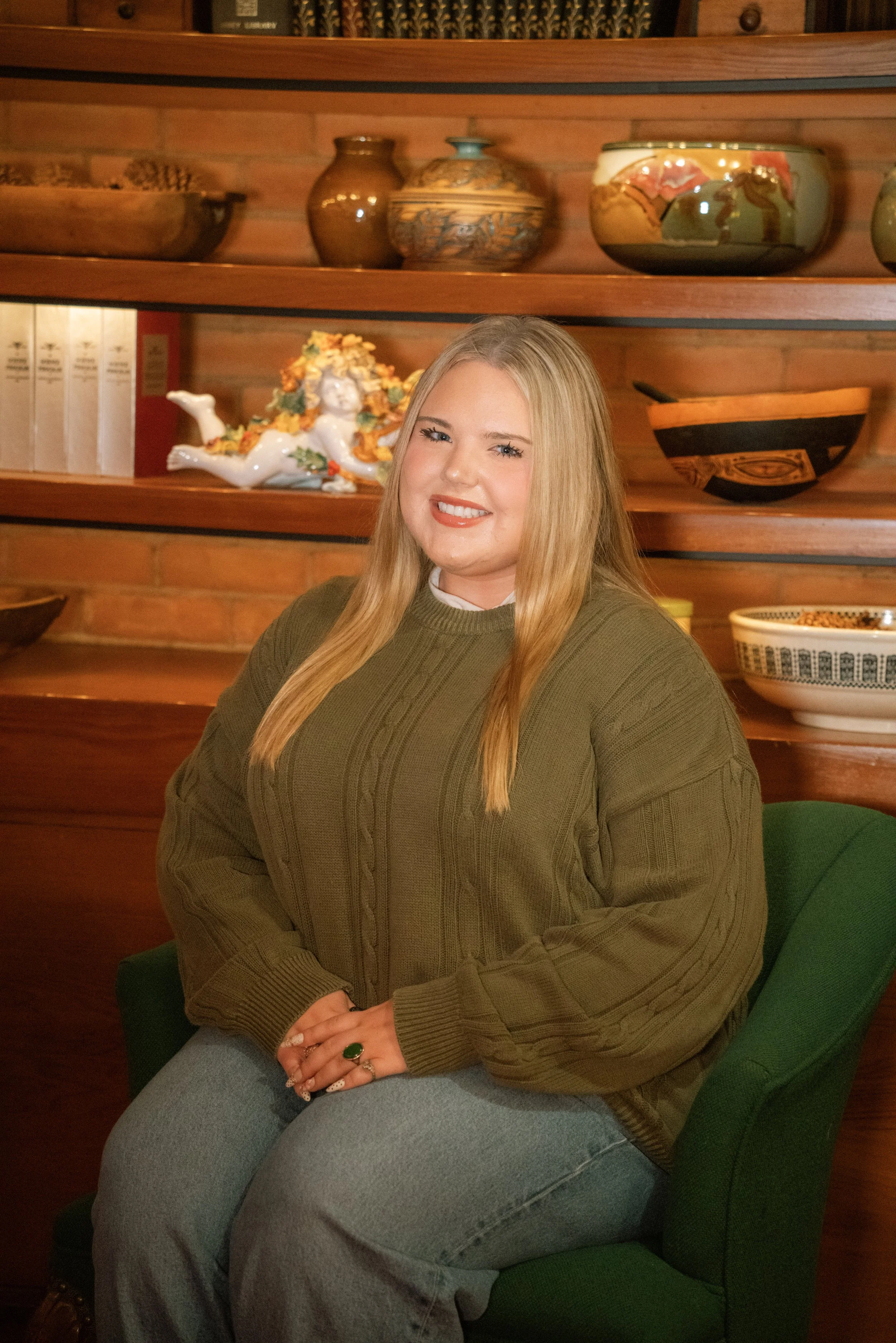 A young woman with long blonde hair sitting on a green cushioned chair in front of wooden shelves with decorative vases and ceramics, smiling at the camera.