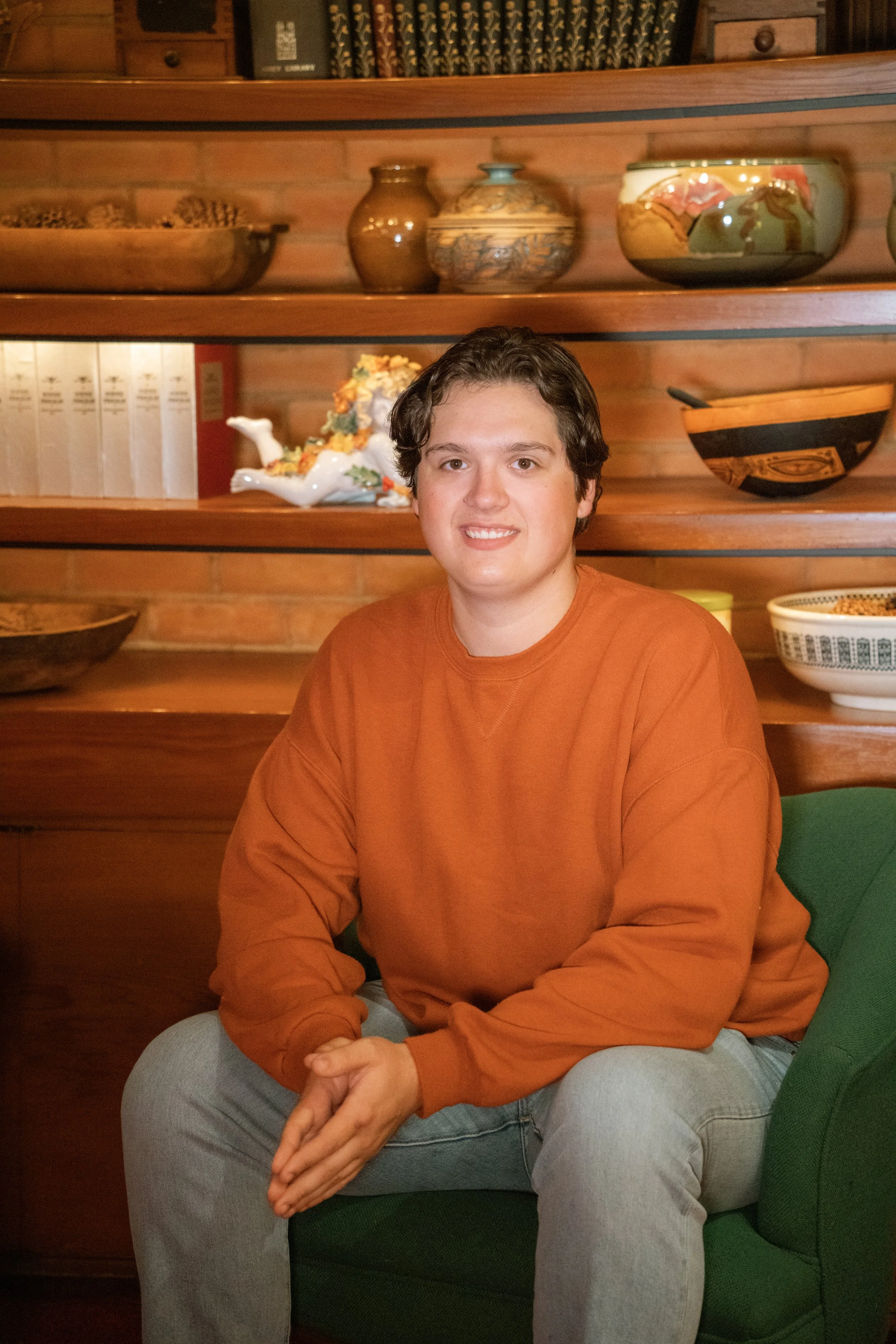 A young man with dark, wavy hair and wearing an orange long sleeve shirt is sitting in front of wooden shelves with decorative bowls and vases in the background.