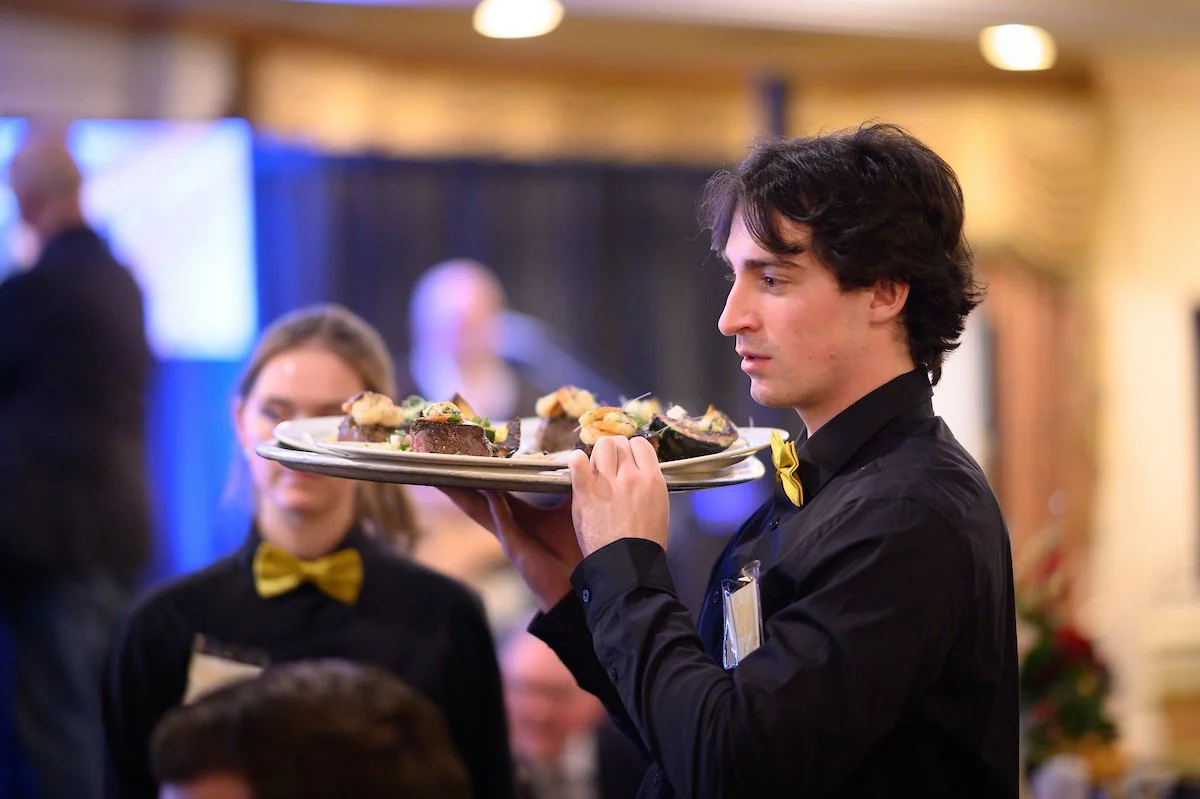 A waiter in a black shirt and yellow bow tie serving a tray of food at a restaurant or event.