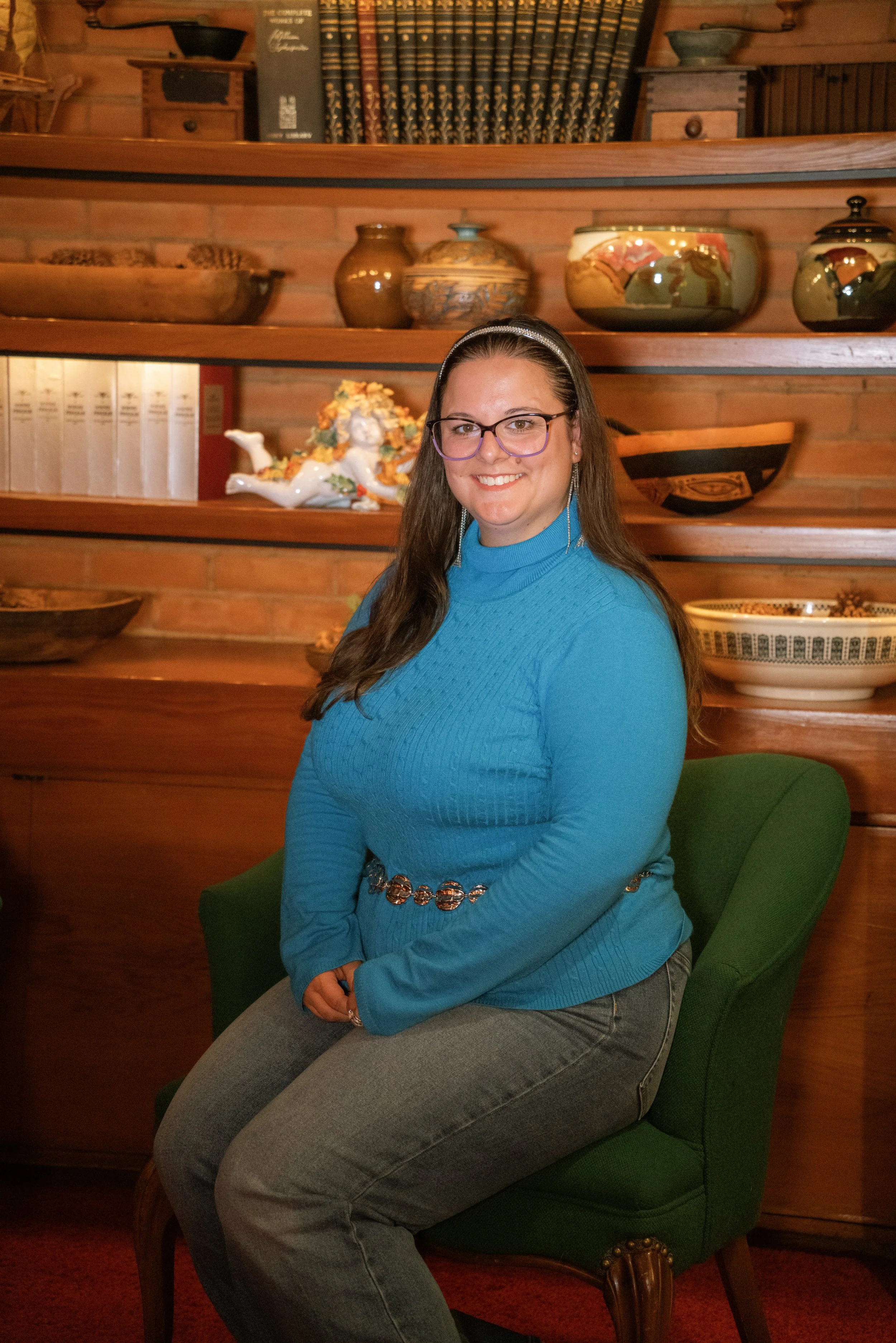 A woman with long dark hair, wearing glasses, a bright blue turtleneck sweater, and gray jeans, sitting on a green upholstered chair in a room with wooden walls. She is smiling at the camera with a backdrop of shelves filled with decorative pottery, books, and a small porcelain figure of an angel.