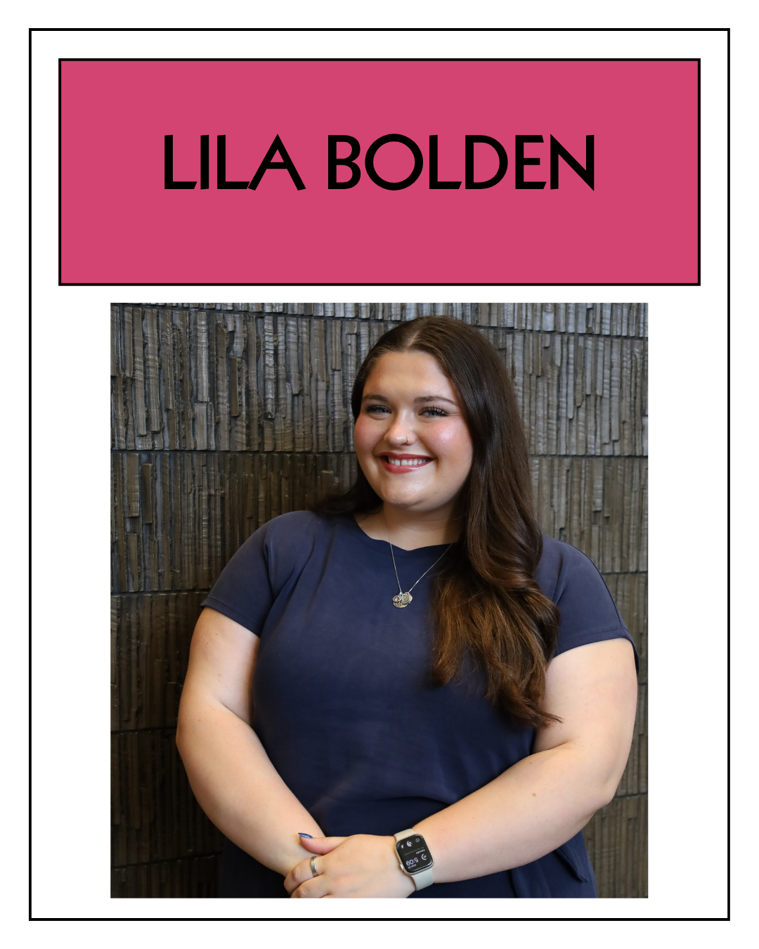 Young woman with long dark hair, wearing a navy blue shirt, smiling, standing with arms crossed in front of a textured wall.