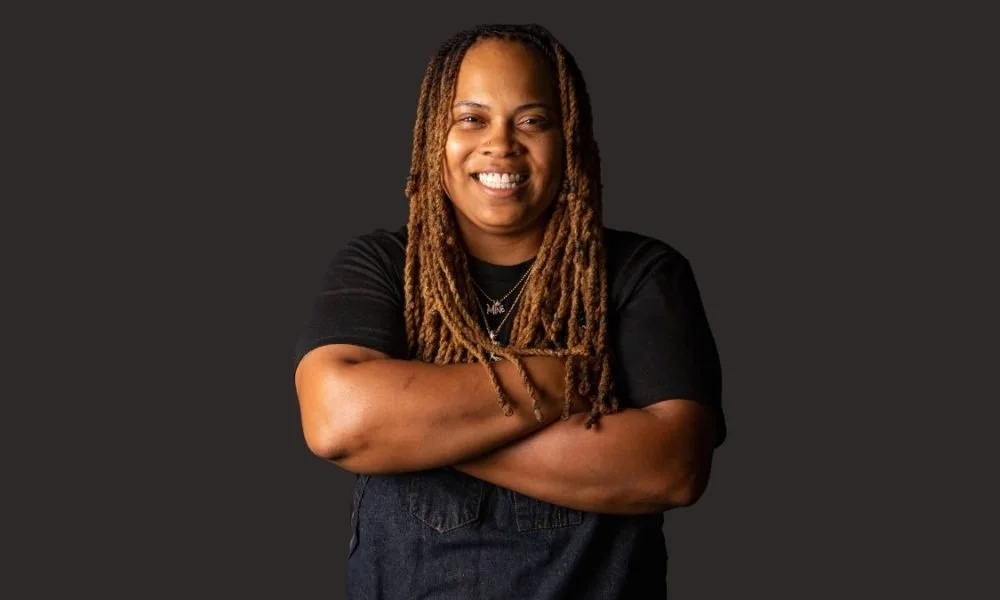 A woman with long dreadlocks smiling with arms crossed, wearing a black T-shirt and jewelry, on a dark background.