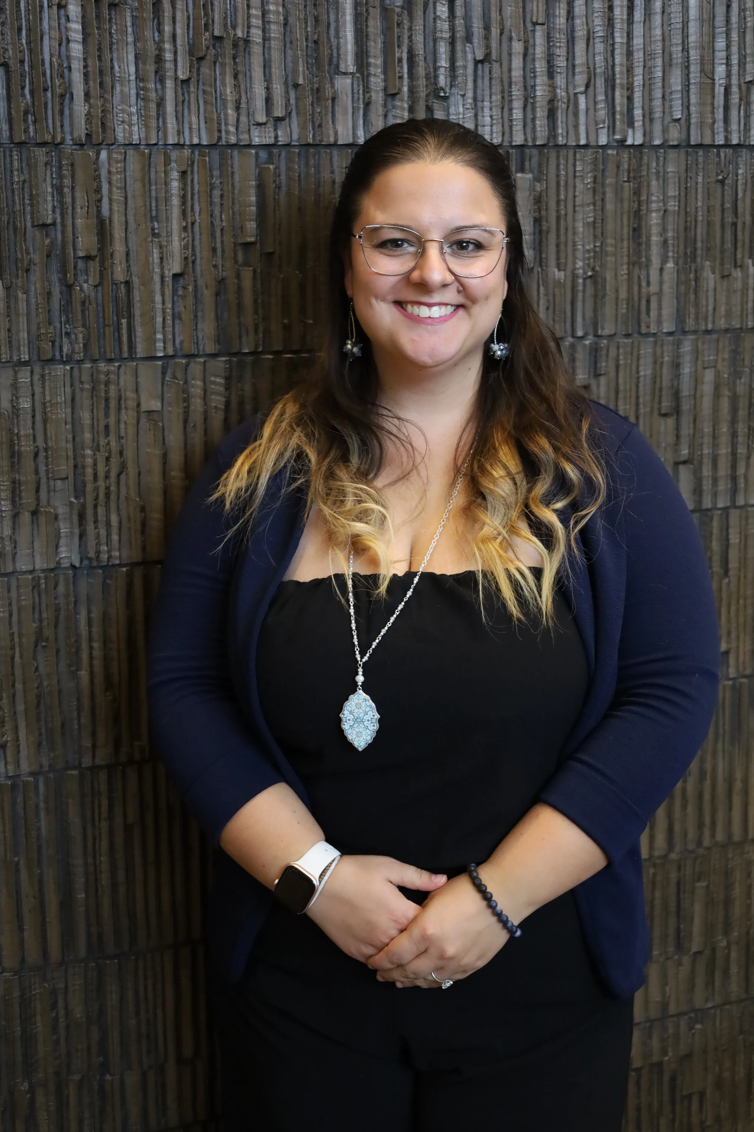 A woman with glasses, long wavy hair, and earrings, standing against a textured brown wall, smiling gently.