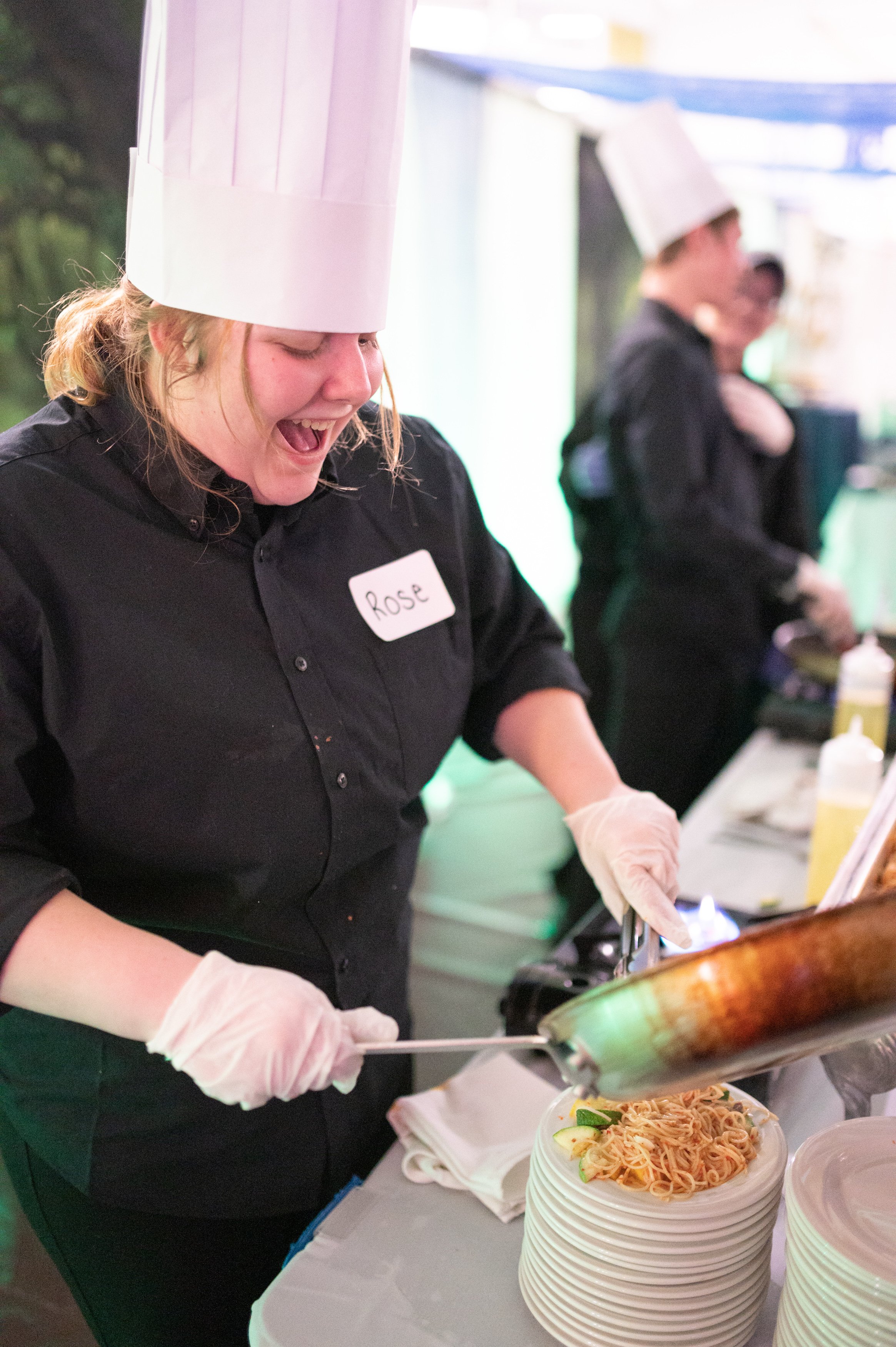 A female chef named Rose serving a plate of spaghetti at a food event, with other chefs working in the background.
