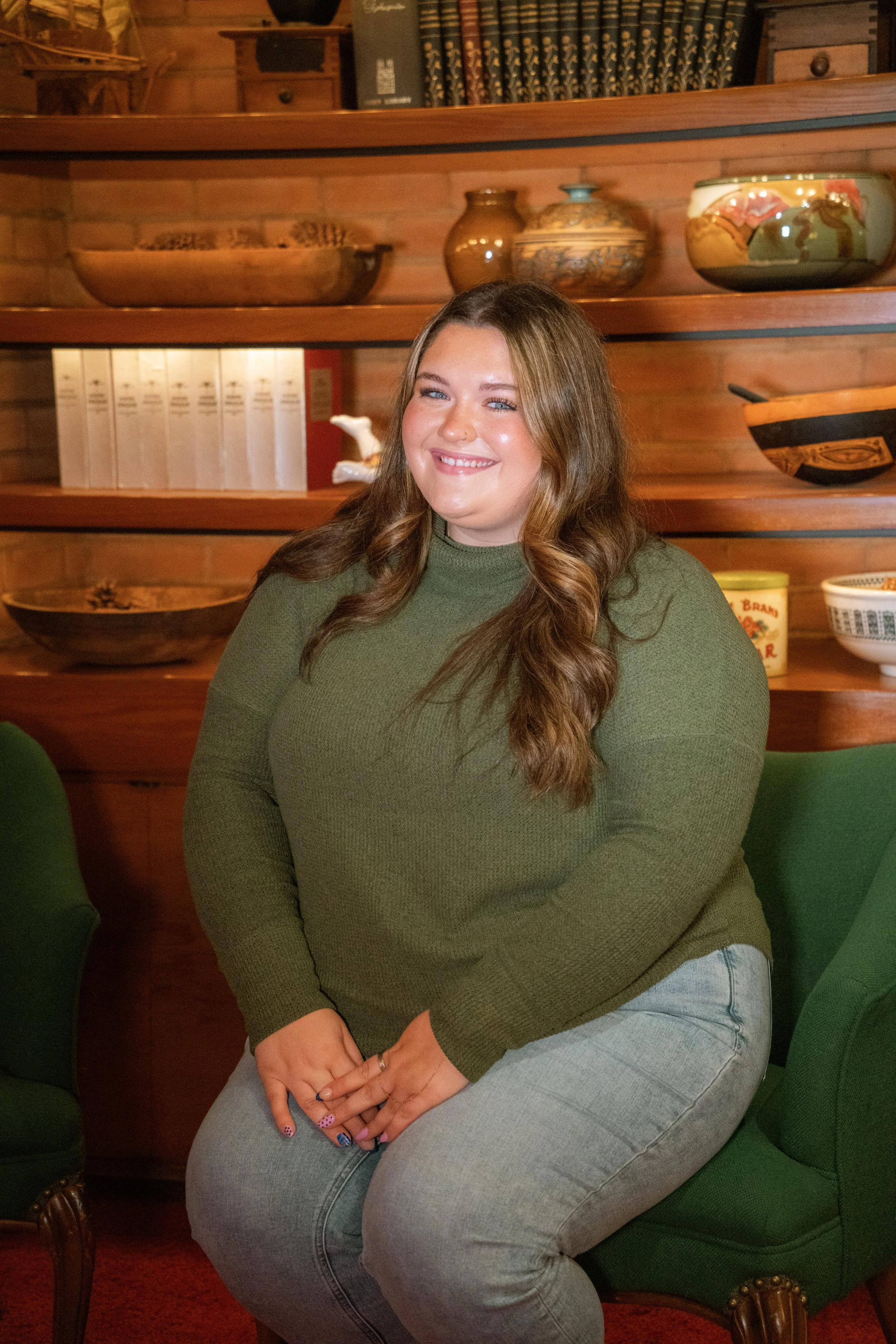 A woman with long, wavy hair, smiling, sitting on a green cushioned chair in front of a wooden wall with decorative bowls and pottery on shelves.