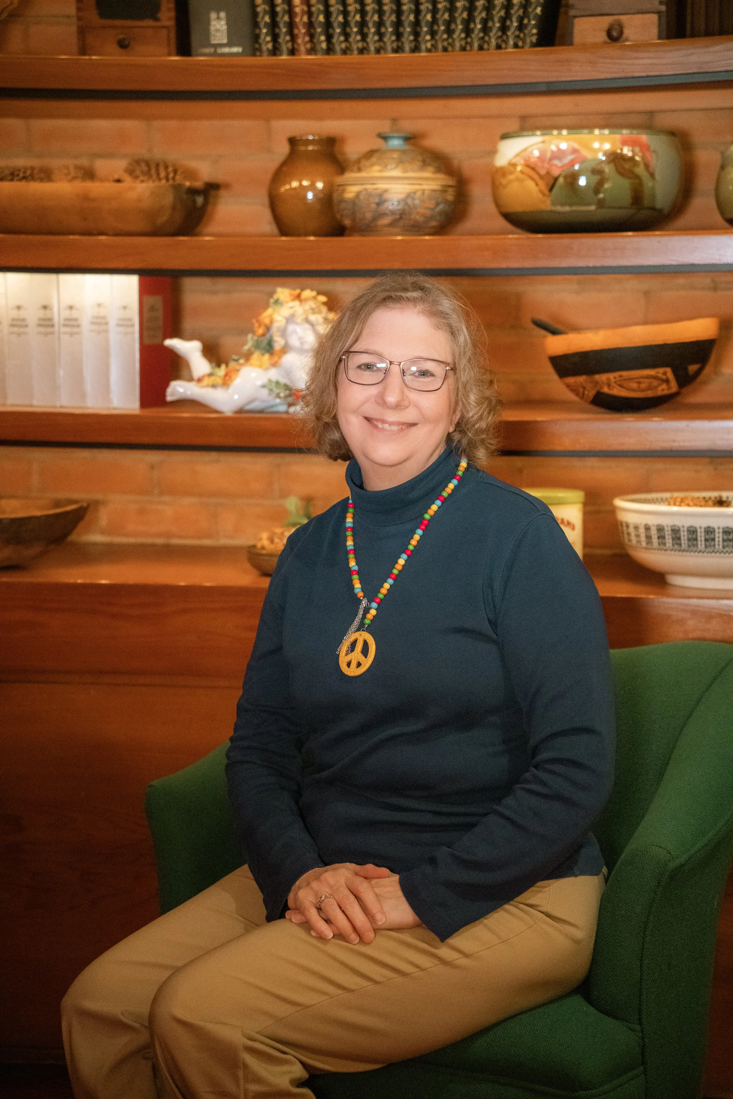 A smiling woman with glasses and blonde hair, wearing a black long-sleeve shirt and tan pants, sitting in a green chair in front of wooden shelves with vases and ceramic sculptures.