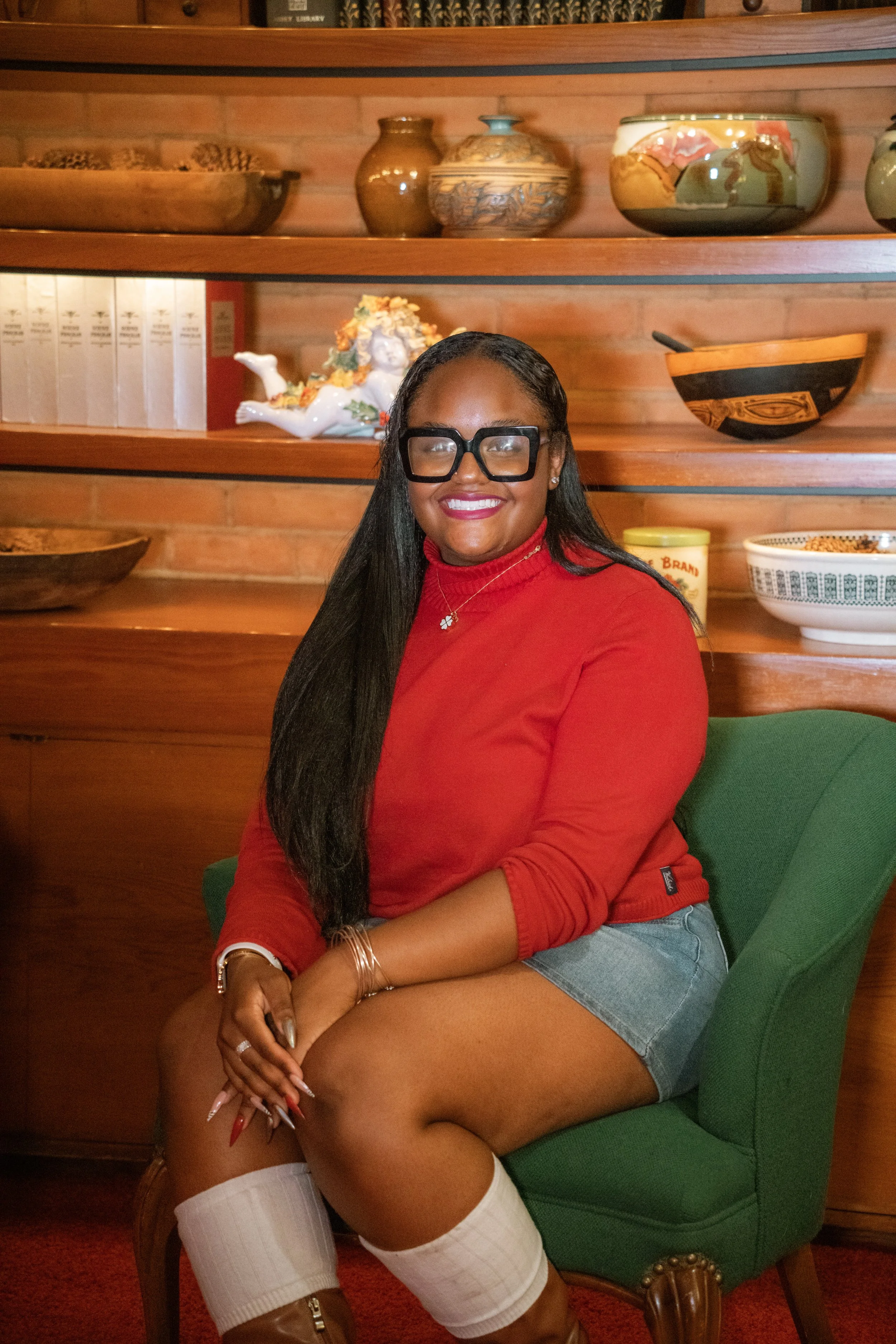 A woman sitting on a green chair in front of a wooden shelf with ceramics and decorative objects behind her. She is wearing glasses, a red long-sleeve shirt, denim shorts, white thigh-high stockings, and brown boots. She has long black hair and is smiling.
