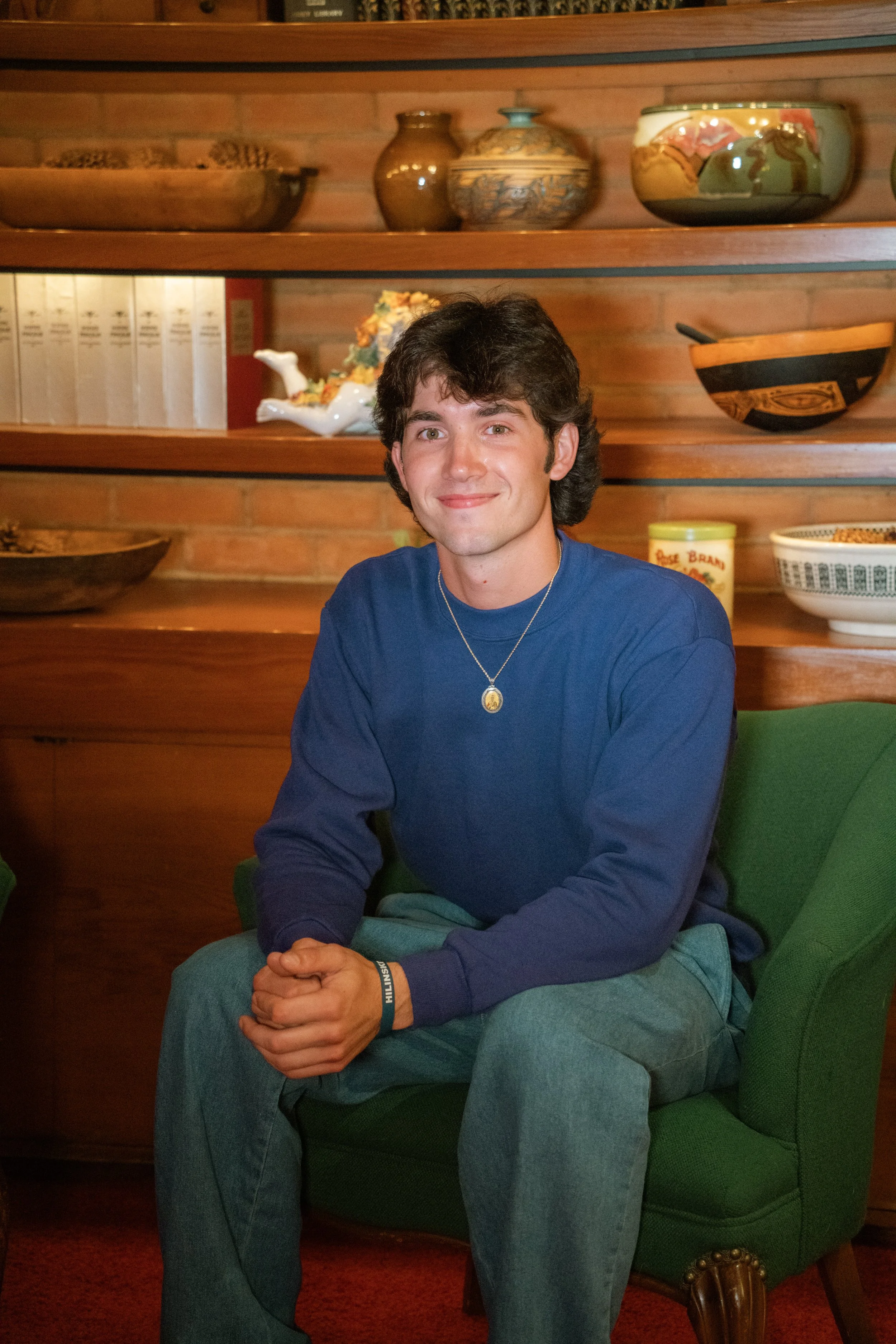 A young man with dark hair and a friendly smile, sitting in a green chair in front of a wooden shelf with decorative bowls and vases, wearing a blue long-sleeve shirt and jeans.