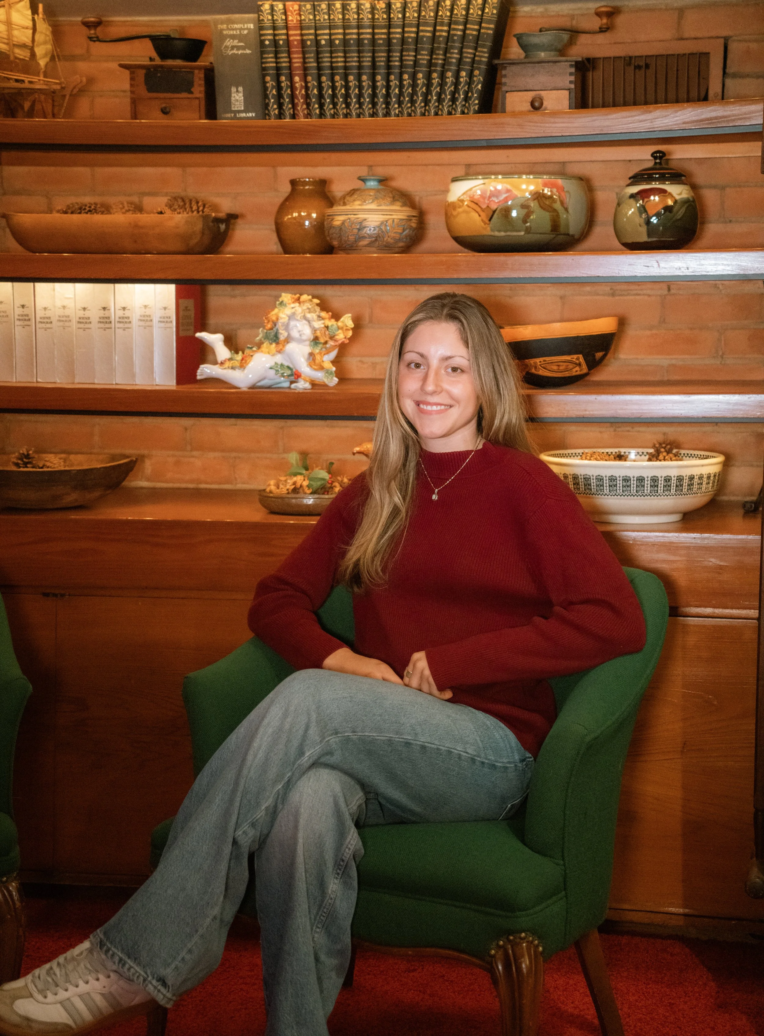 A young woman with long light brown hair, wearing a red sweater and gray jeans, is sitting and smiling in a green armchair in front of wooden shelves with decorative pottery and books. The shelves are against a brick wall.