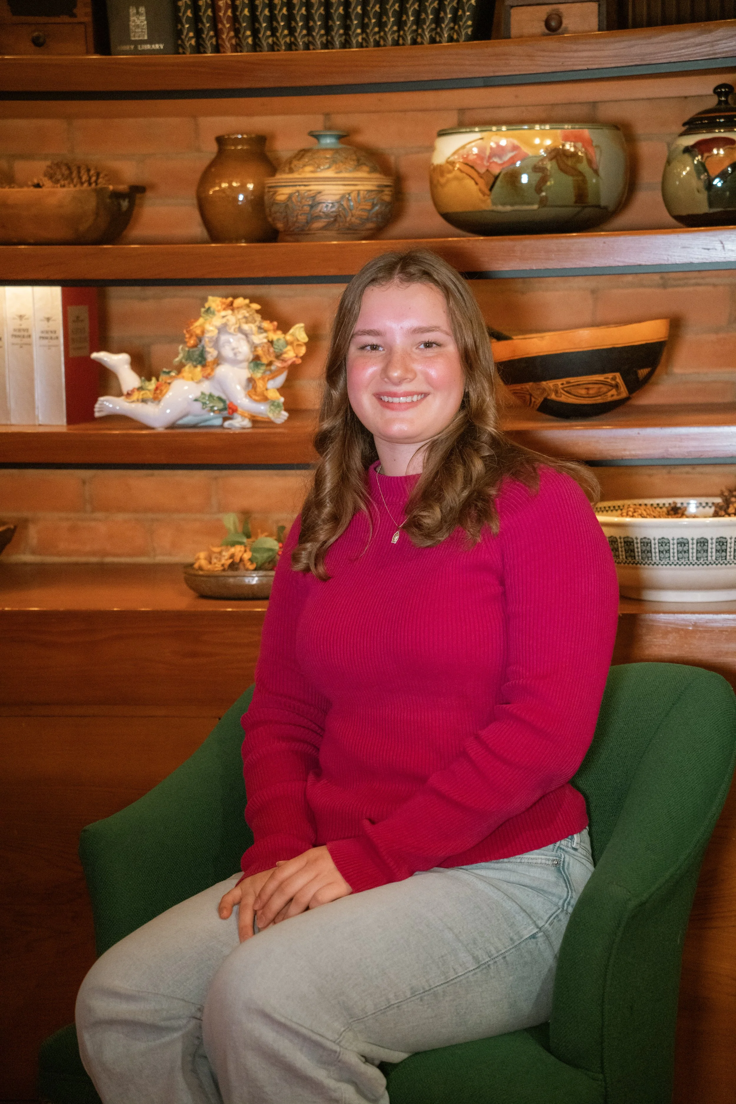 A young woman with long wavy hair, wearing a pink sweater and light-colored pants, sitting on a green chair in front of wooden shelves with decorative pottery and books, smiling at the camera.