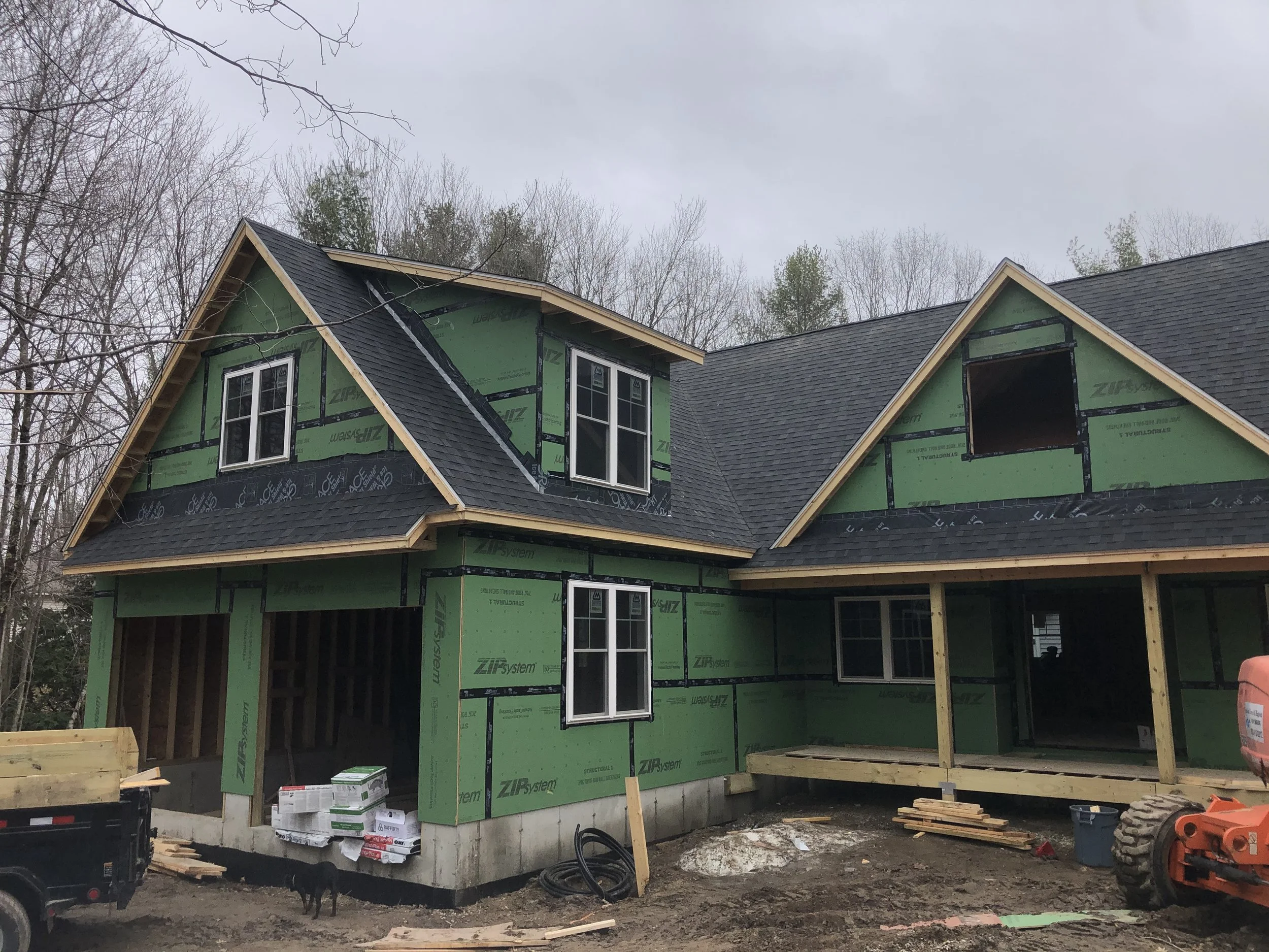 House under construction with green weather-resistant sheathing, black roofing, and scaffolding. Construction materials and equipment are visible outside.