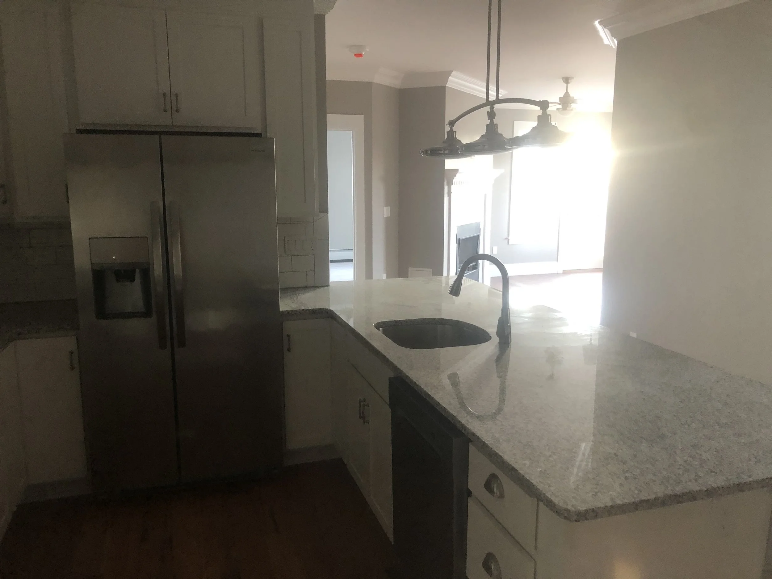 Kitchen with a granite countertop, stainless steel refrigerator, sink, and hanging light fixtures, bright sunlight streaming in from the windows.