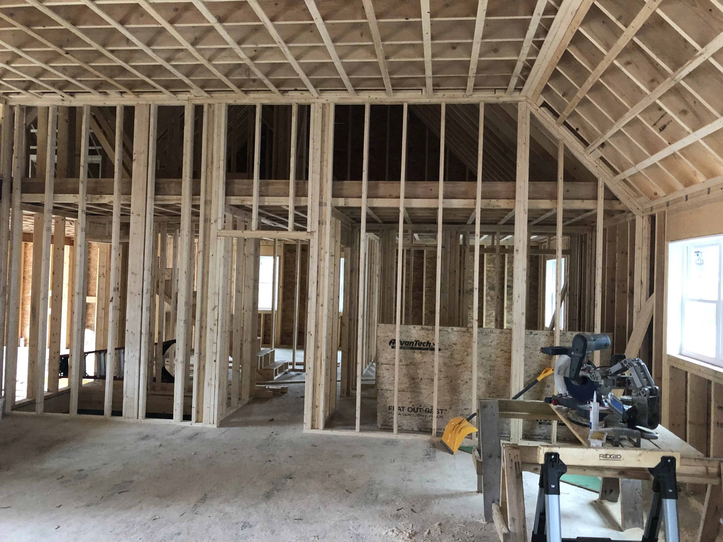 Interior view of a house under construction, with exposed wooden framing and a sloped ceiling. Construction tools are visible on a makeshift workbench in the foreground, and windows are installed on the right side.