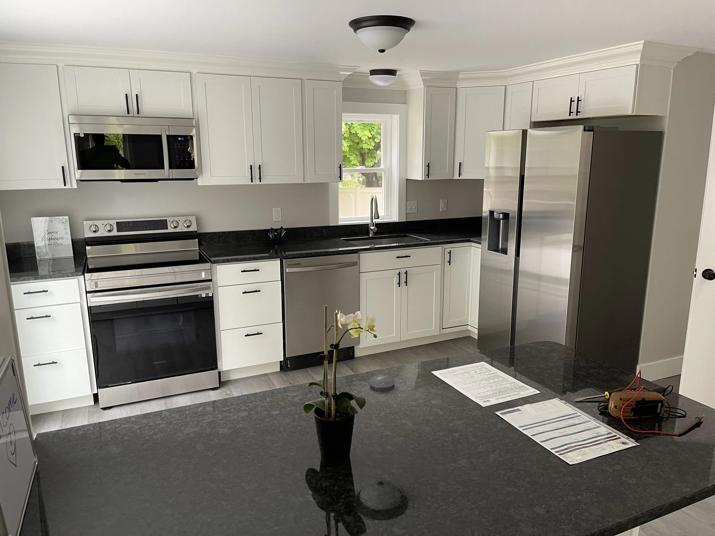 Modern kitchen with white cabinets, black countertops, stainless steel appliances, and a window above the sink. There is a potted orchid on the kitchen island and some papers and electronic tools on the counter.