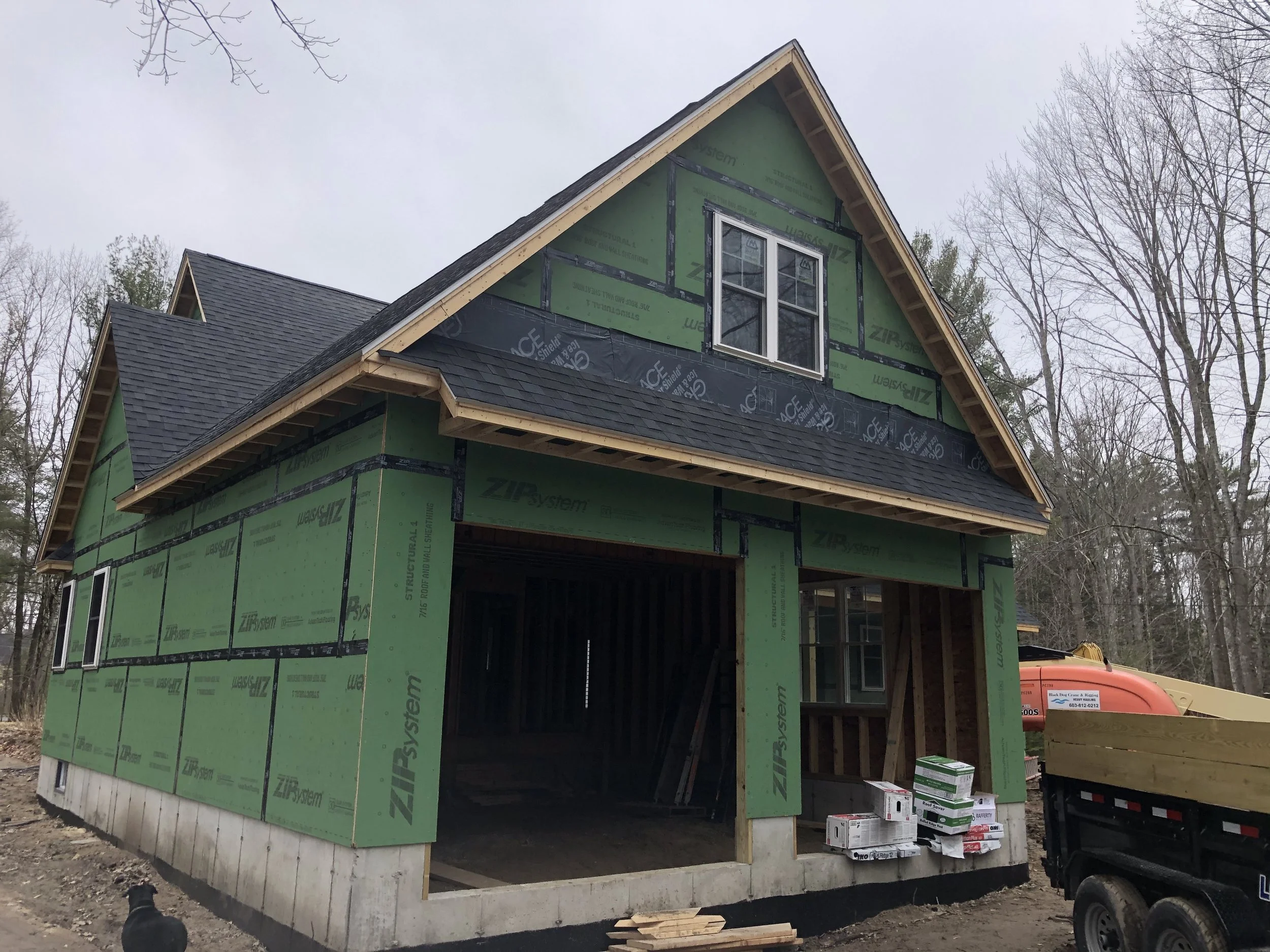 A house under construction with green sheathing, a black roof, and an open garage area, surrounded by trees and construction equipment.