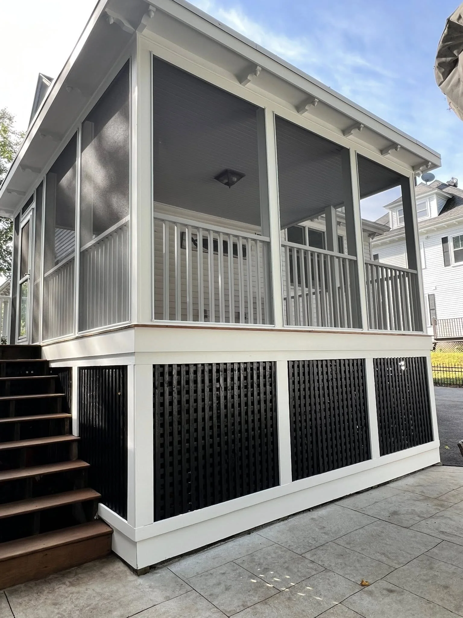 A screened porch with black and white wooden railings and stairs, attached to a house with a decorative ceiling and outdoor lighting, under a partly cloudy sky.