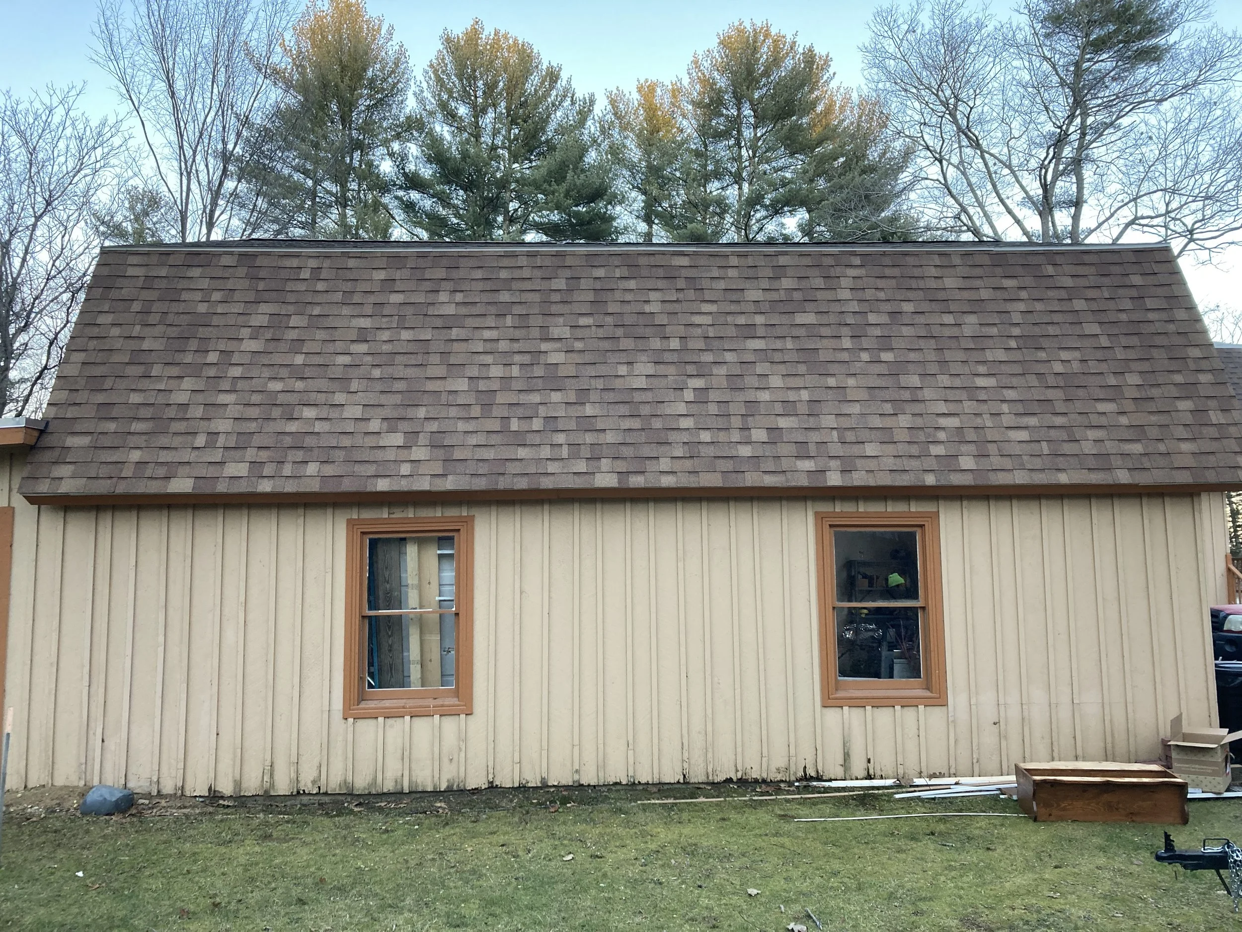 A house with a brown shingled roof, beige vertical siding, and two wooden-framed windows. There is a grassy yard in front, with some construction materials and boxes visible near the house.