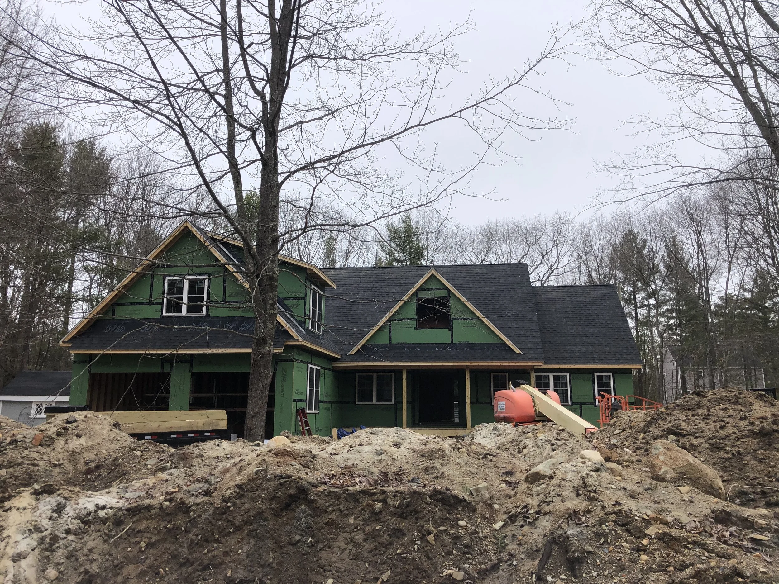 A house under construction with green sheathing, black roof, and a large pile of dirt and construction equipment in the foreground.