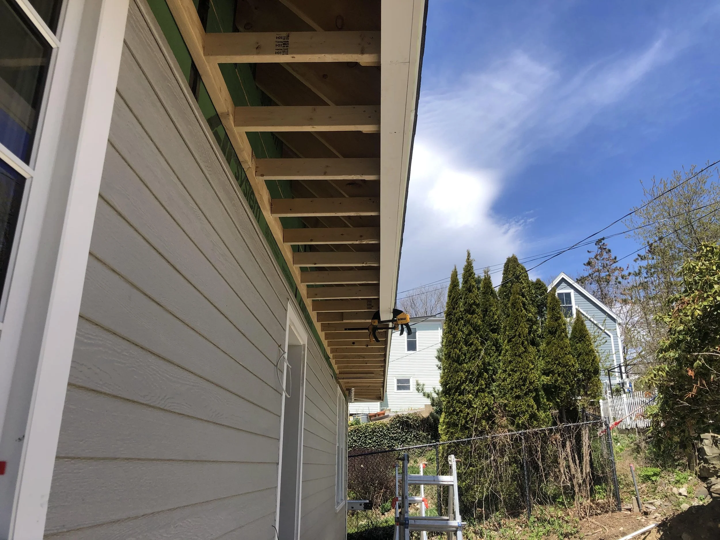 Side of a house with beige siding, under construction, with an overhang roof frame, ladder in the yard, surrounded by trees and neighboring houses under a partly cloudy sky.