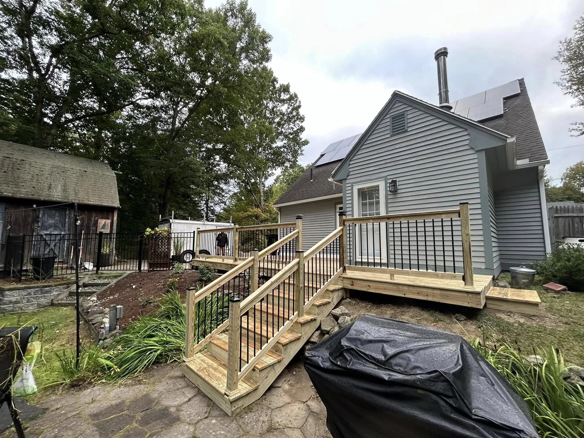 Backyard with new wooden deck under construction, attached to a gray house with a chimney and solar panels, surrounded by trees, with a black grill covered in the foreground.