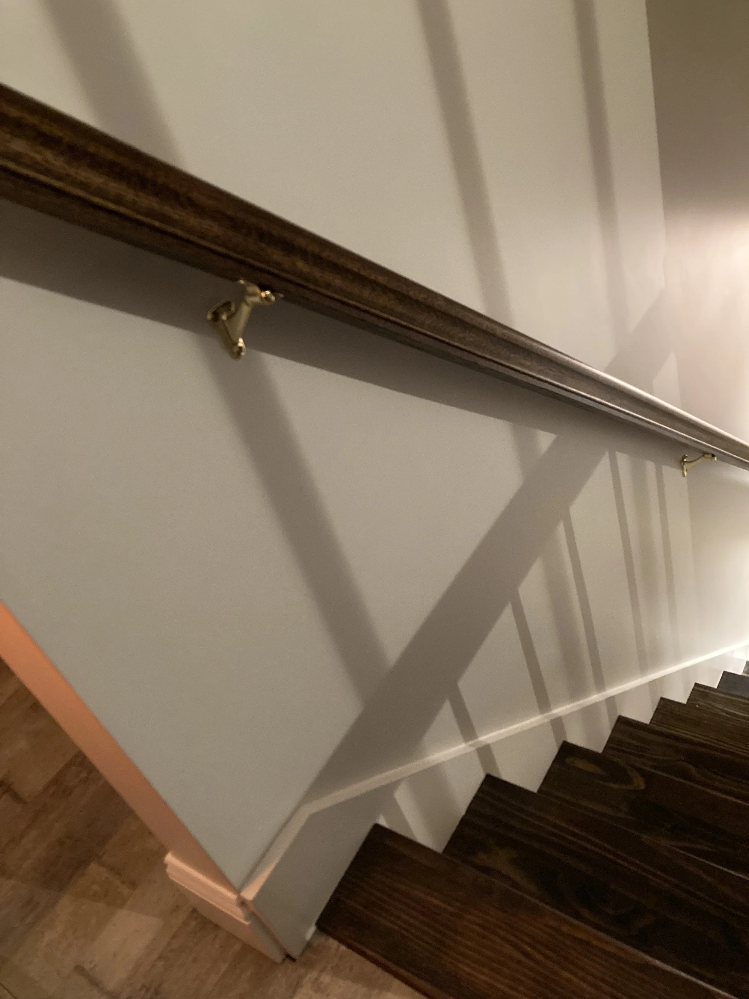 Interior view of a staircase with dark wooden steps, a light-colored wall, and a wooden handrail supported by golden brackets.