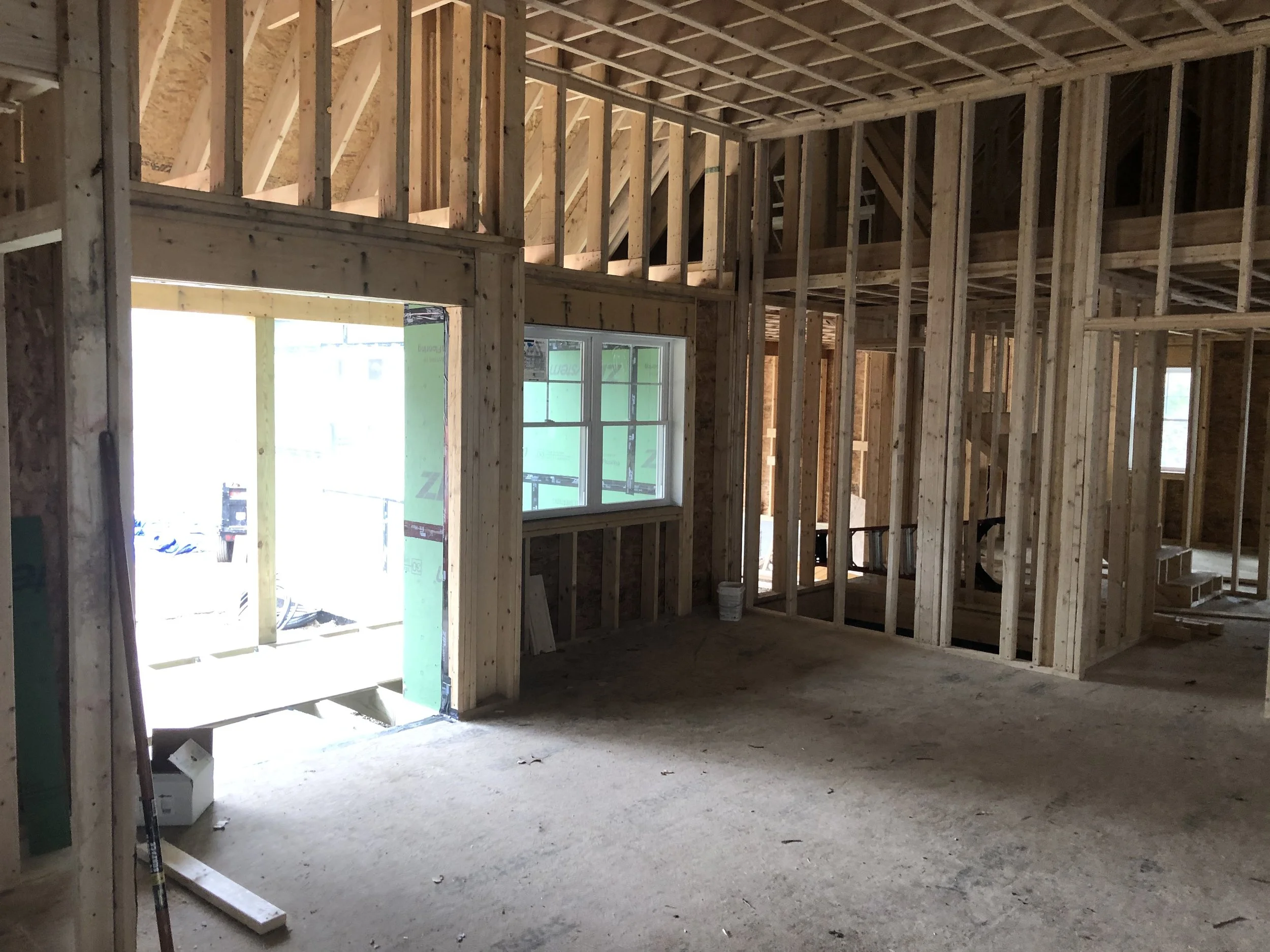 Interior of a house under construction with exposed wooden framing, stairs, and a window, unfinished flooring.