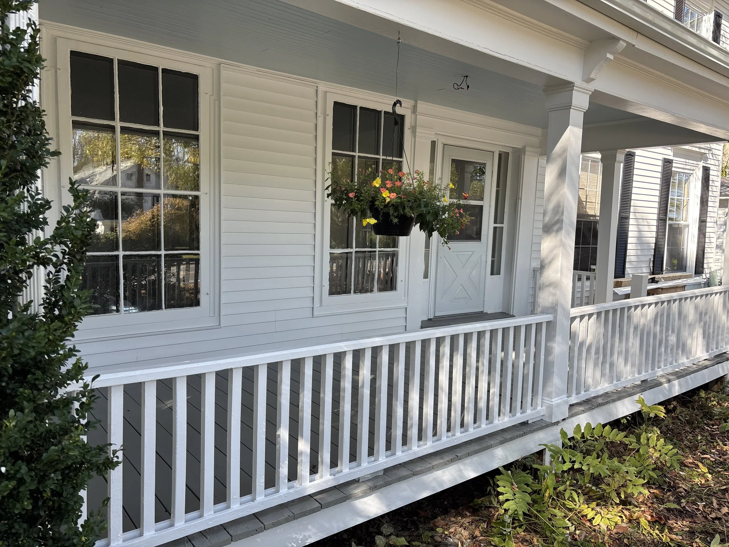 White house porch with white railing, hanging flower basket, and front door with glass panels, surrounded by windows with black shutters and shrubbery.