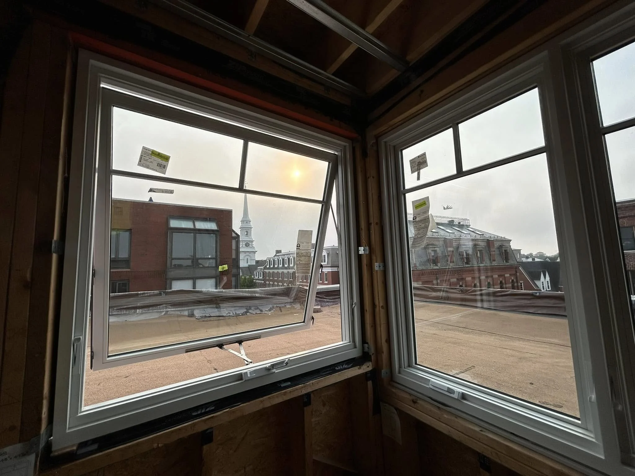 Interior view of a room under construction with three large windows, one of which is open, showing a cityscape with buildings, a church steeple, and a cloudy sky with the sun partially visible.