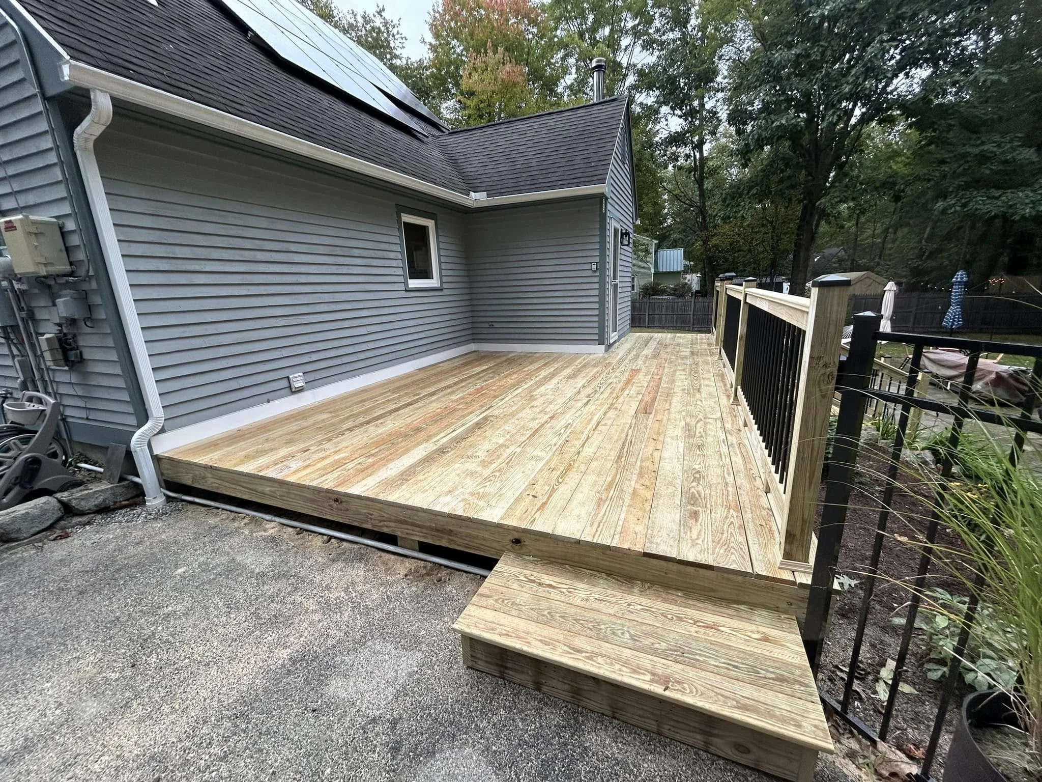 Newly built wooden deck attached to a gray house with gray siding in a backyard, with steps leading up to the deck and a black fence surrounding part of the yard, with trees and outdoor furniture in the background.