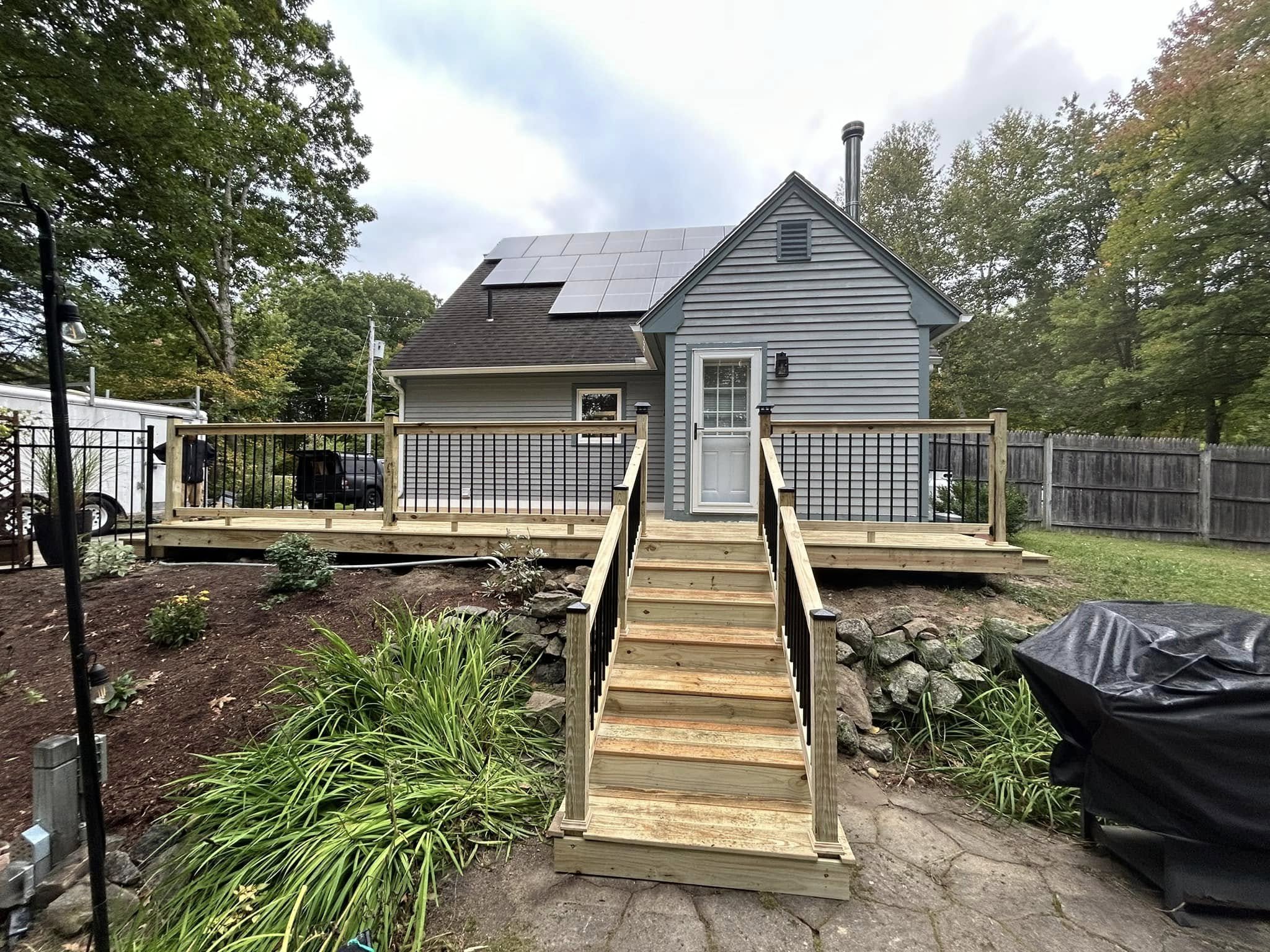 Newly built wooden deck with stairs leading up to a house with solar panels on the roof, surrounded by trees and a fenced yard.