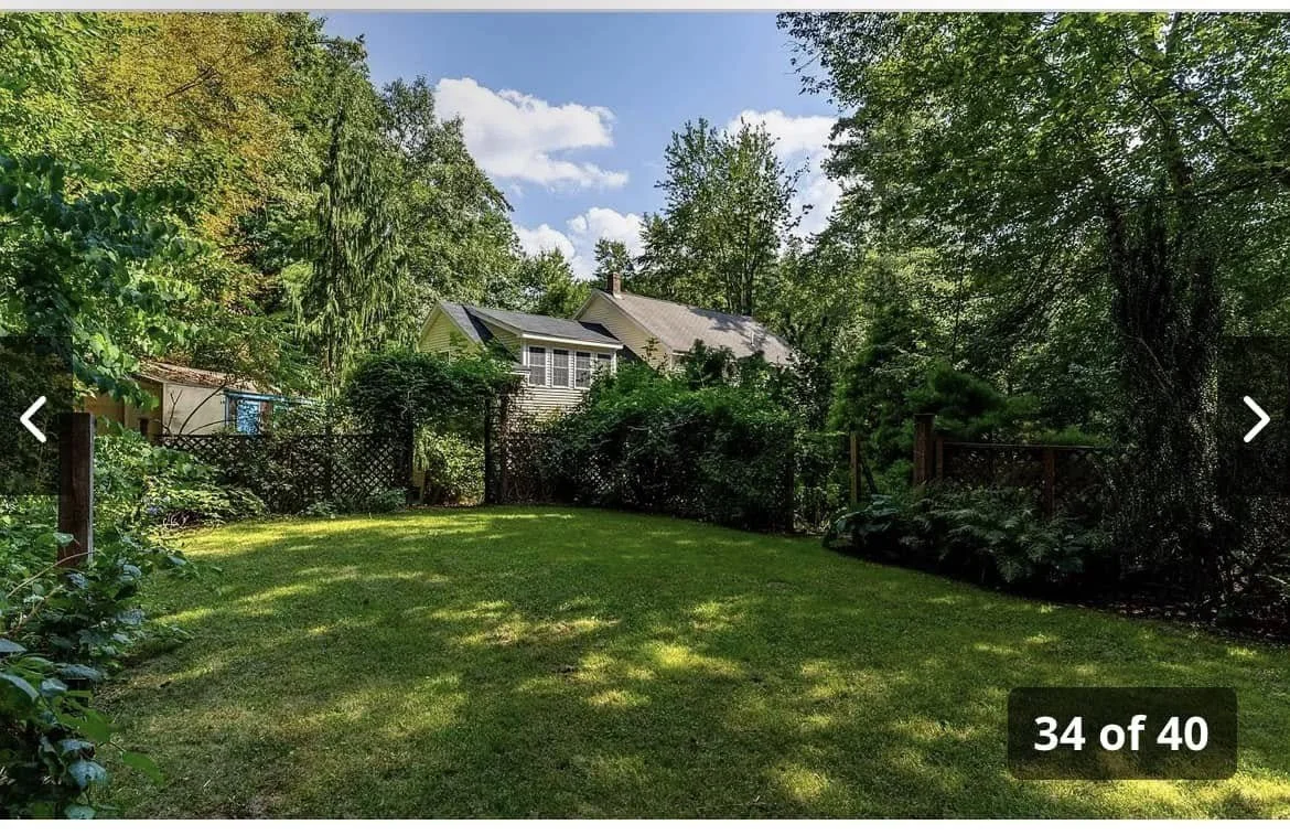 Backyard with a well-maintained grass lawn, surrounded by tall trees and shrubs, with a house partially visible in the background under a blue sky with clouds.